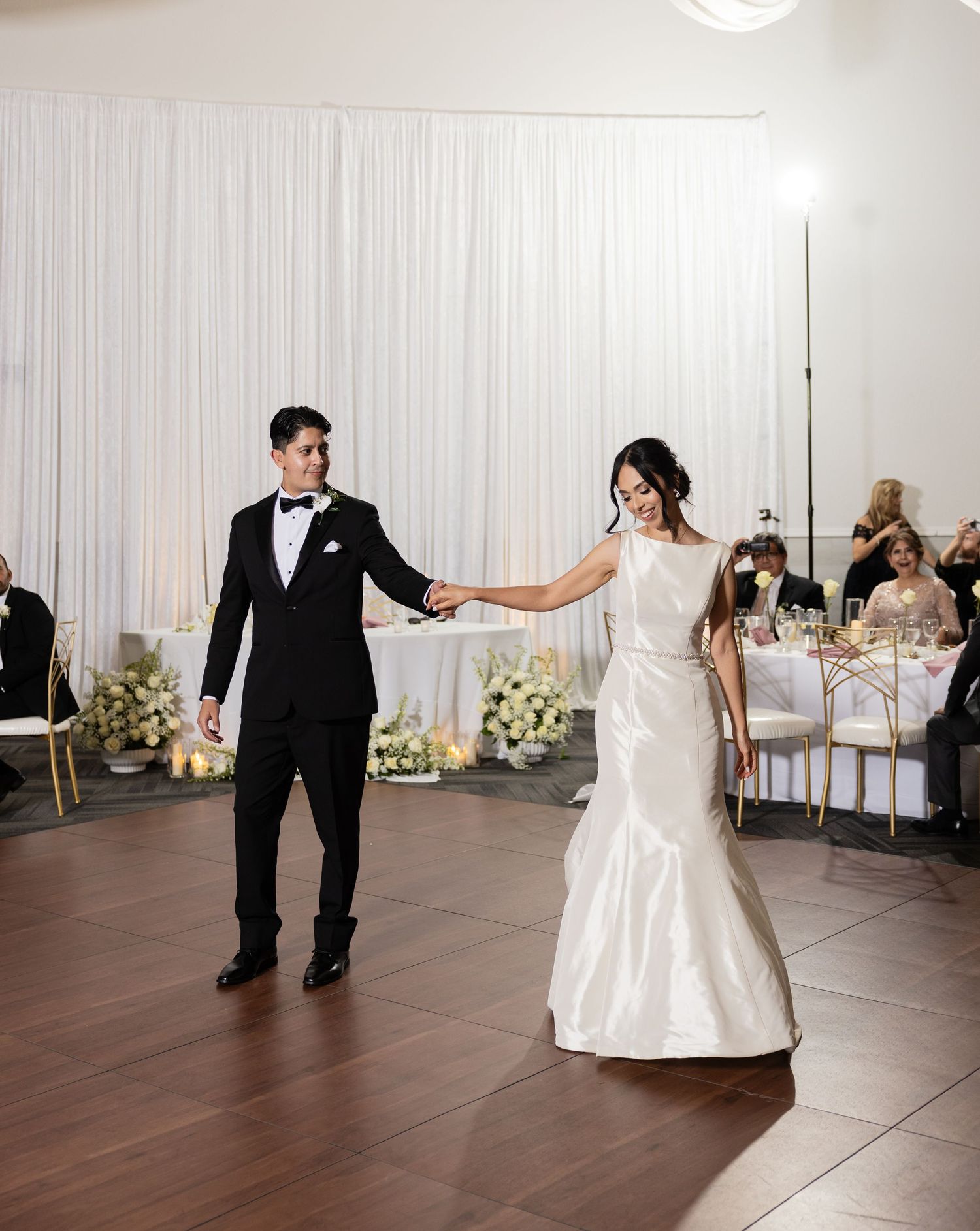 Newlyweds glide across a wooden dance floor during their wedding reception in a white-decorated ballroom.