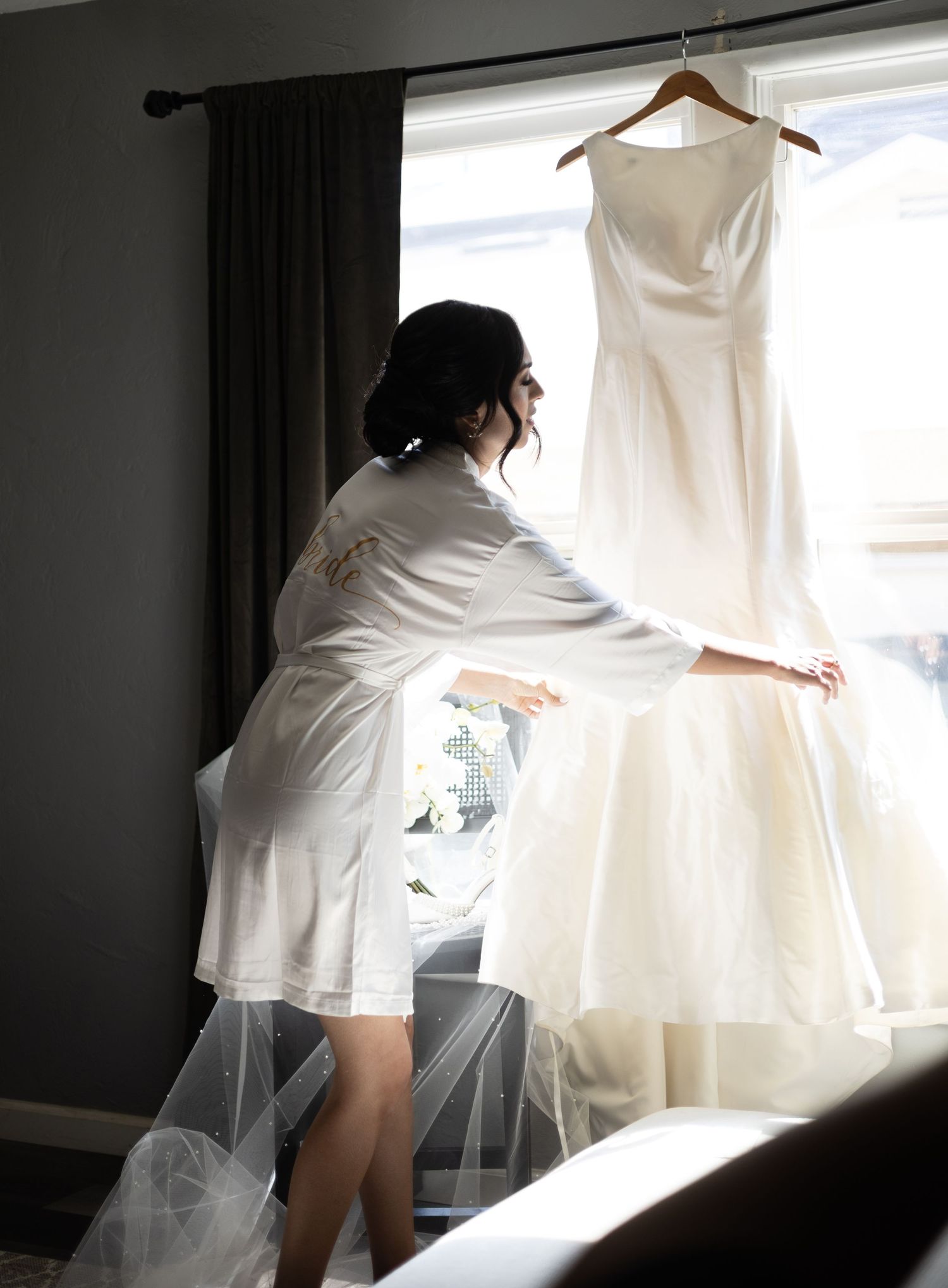 Silhouette of someone in white robe reaching for hanging wedding dress near window.