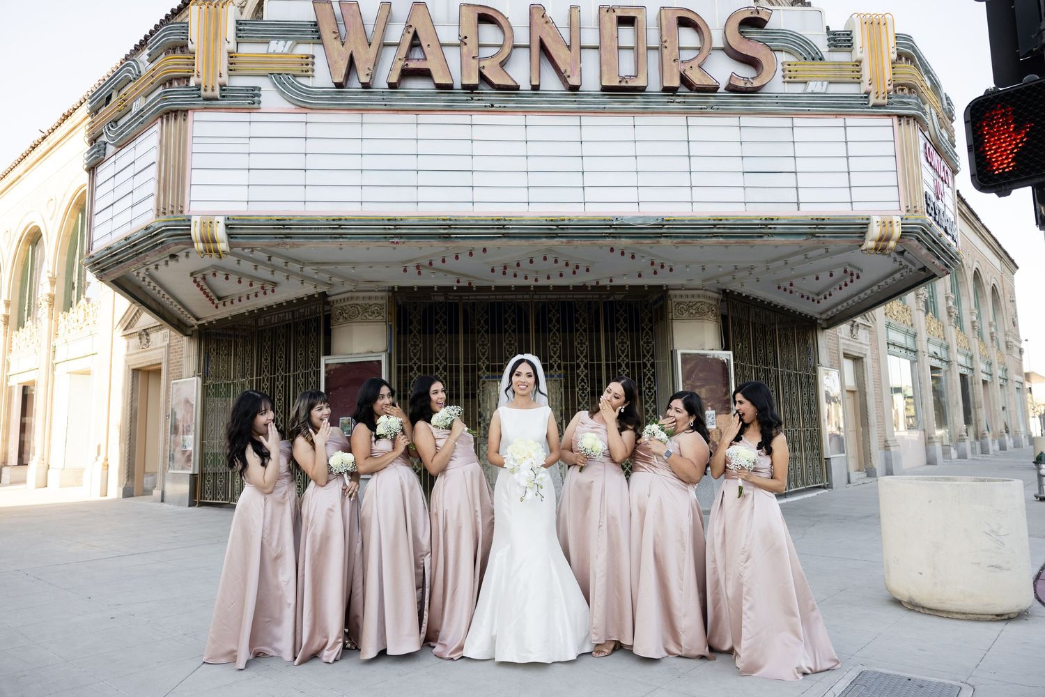 Bridal party in light pink gowns gathered outside vintage theatre marquee.