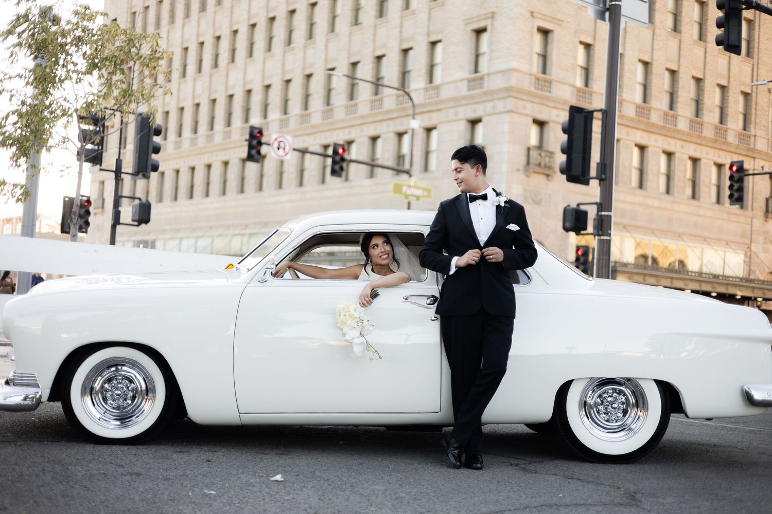 Vintage white car parked in front of urban building with couple in formal attire.
