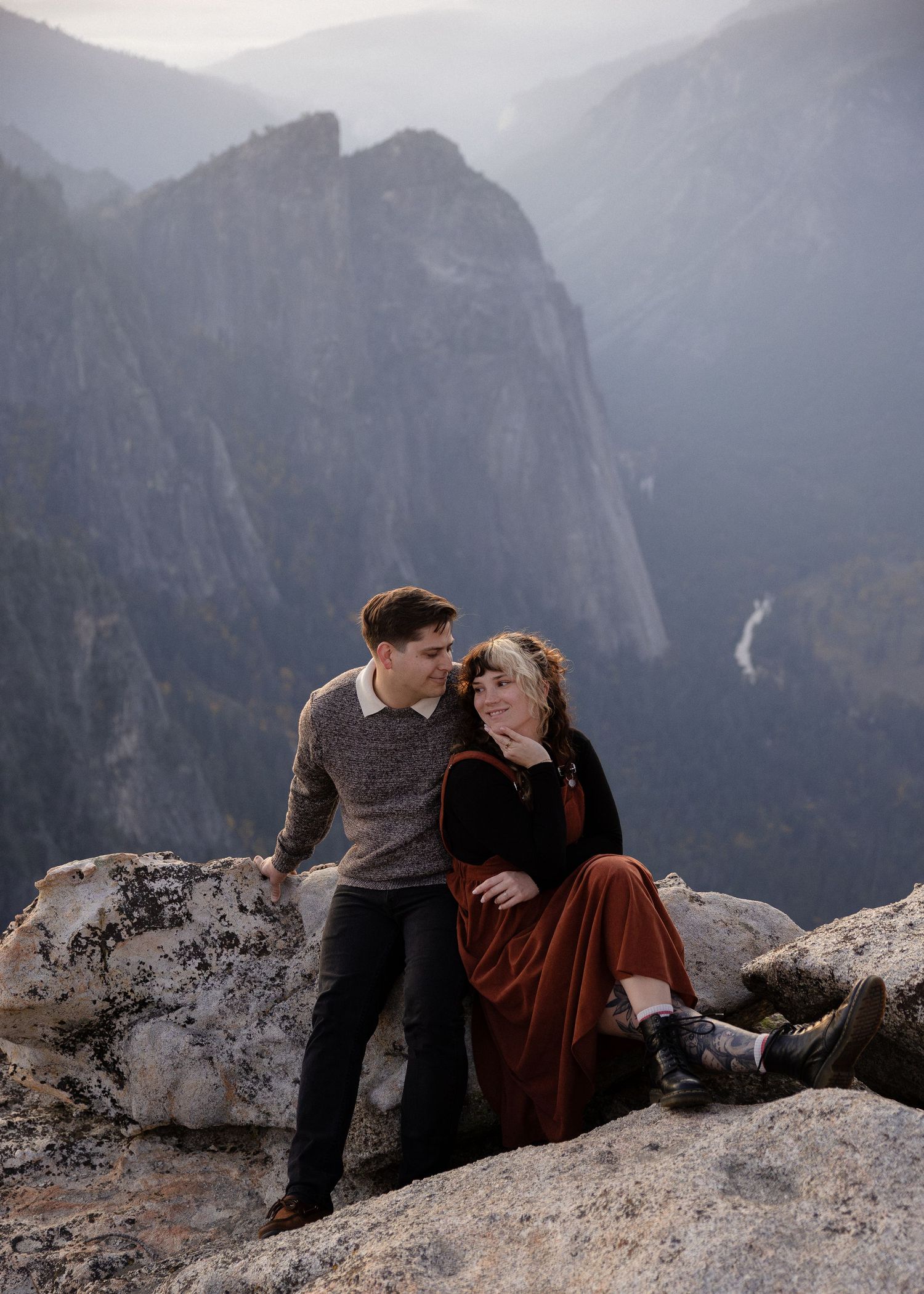 Couple sitting on rocky cliff edge overlooking Yosemite Valley's majestic mountain landscape.