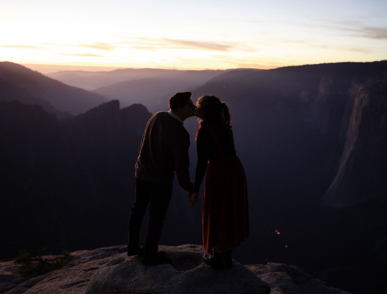 Couple shares intimate moment on mountain cliff edge during sunset.