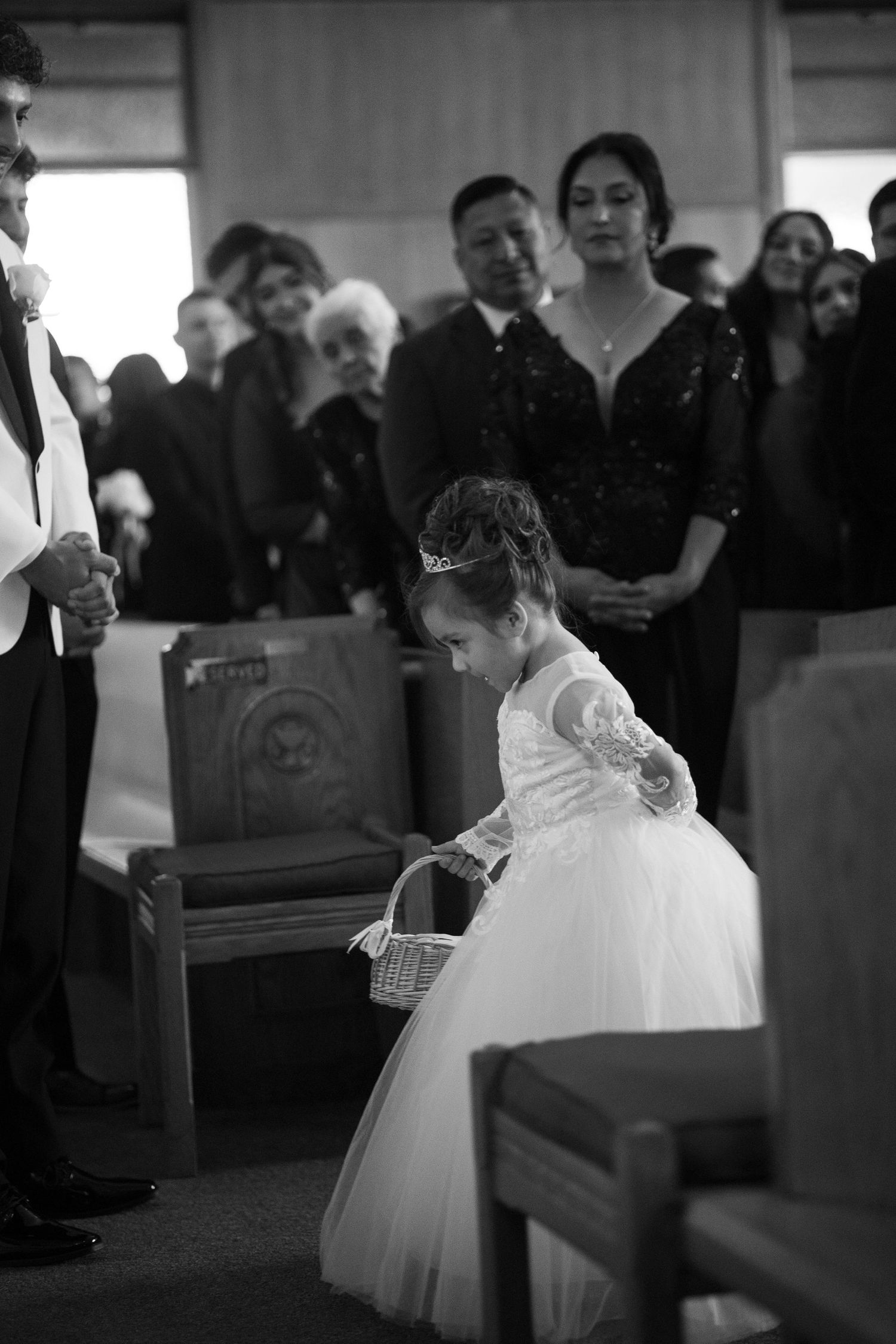 A small flower girl in a white tulle dress carries a basket down the church aisle.