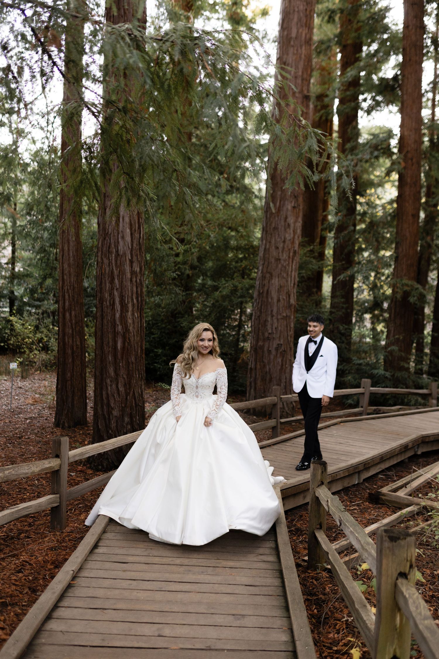 A wedding couple shares a moment on a wooden boardwalk surrounded by tall redwood trees.