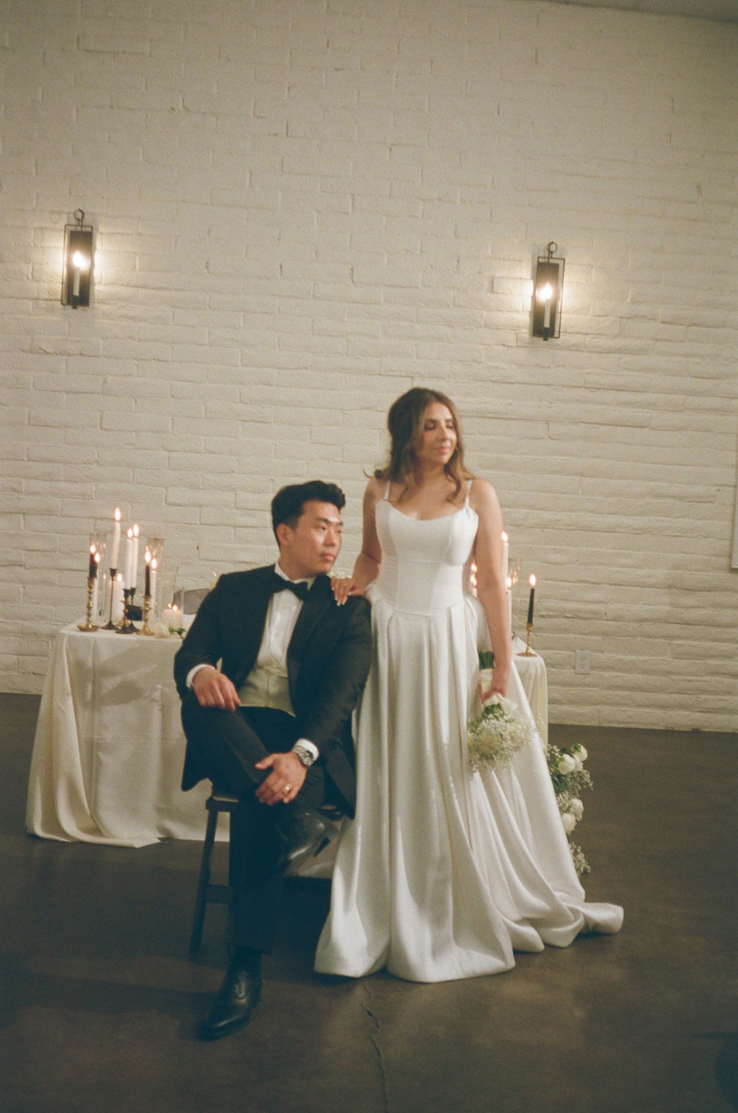 Elegant bride and groom posing at candlelit sweetheart table against white brick wall in wedding venue.
