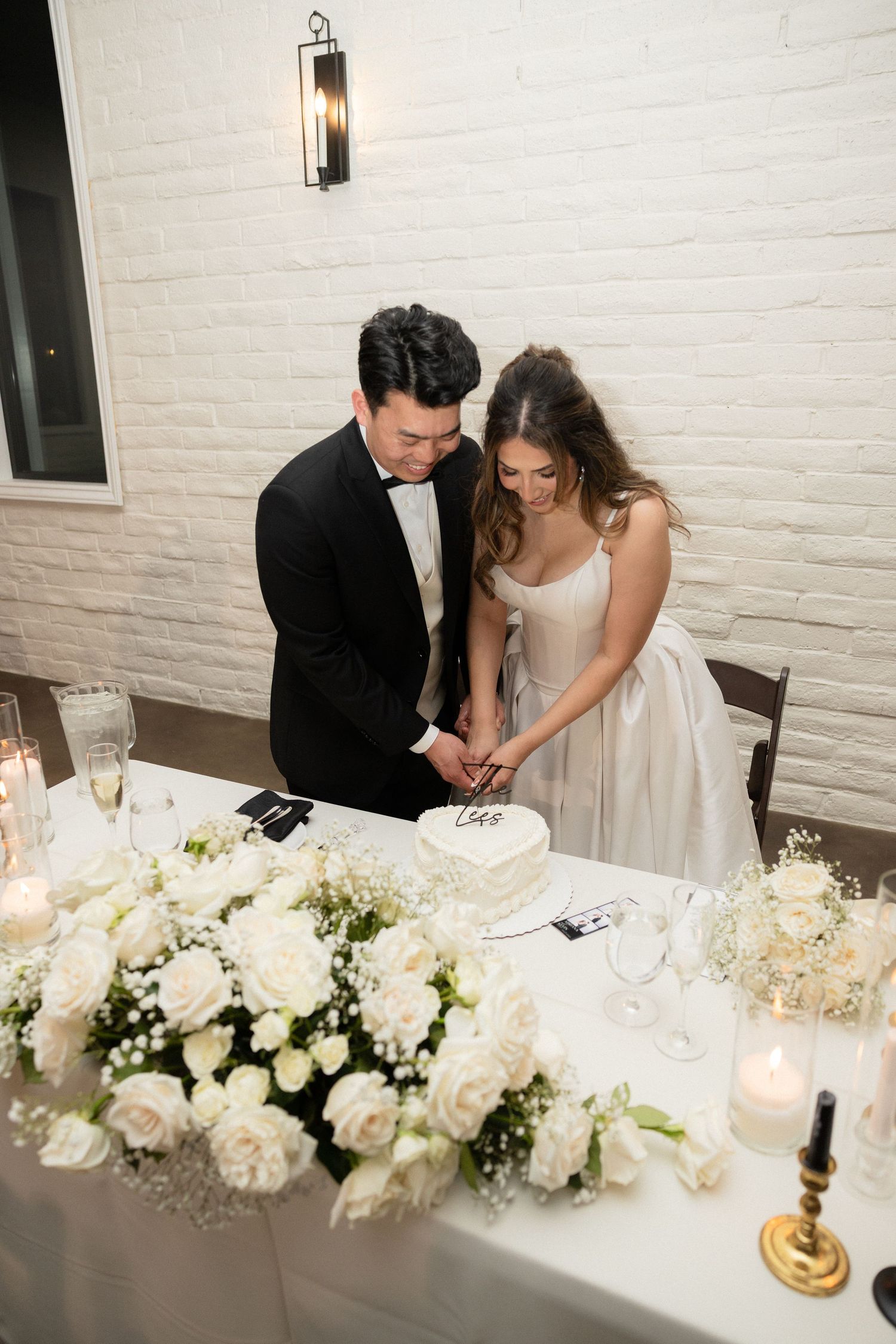 Bride and groom cut heart-shaped wedding cake surrounded by white roses and candles.
