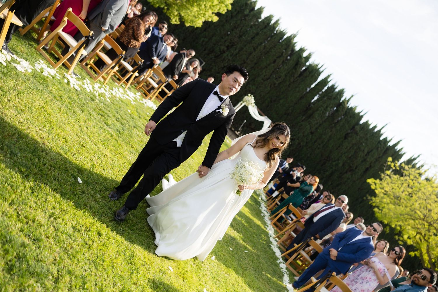 Bride and groom walk hand-in-hand down the aisle at an outdoor wedding ceremony on a sunny day.