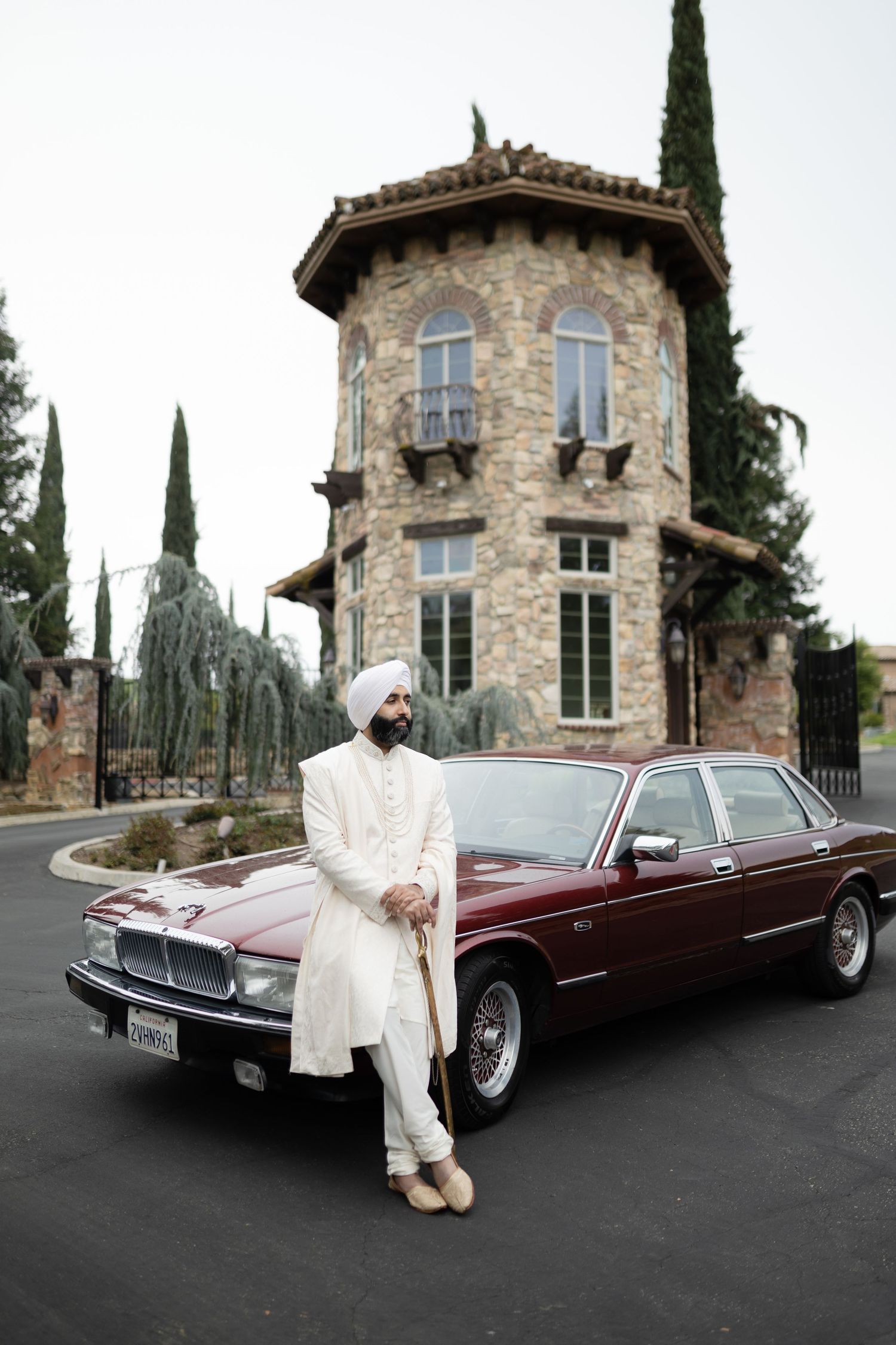 Classic burgundy Lincoln Continental parked in front of a Mediterranean style stone villa with cypress trees.