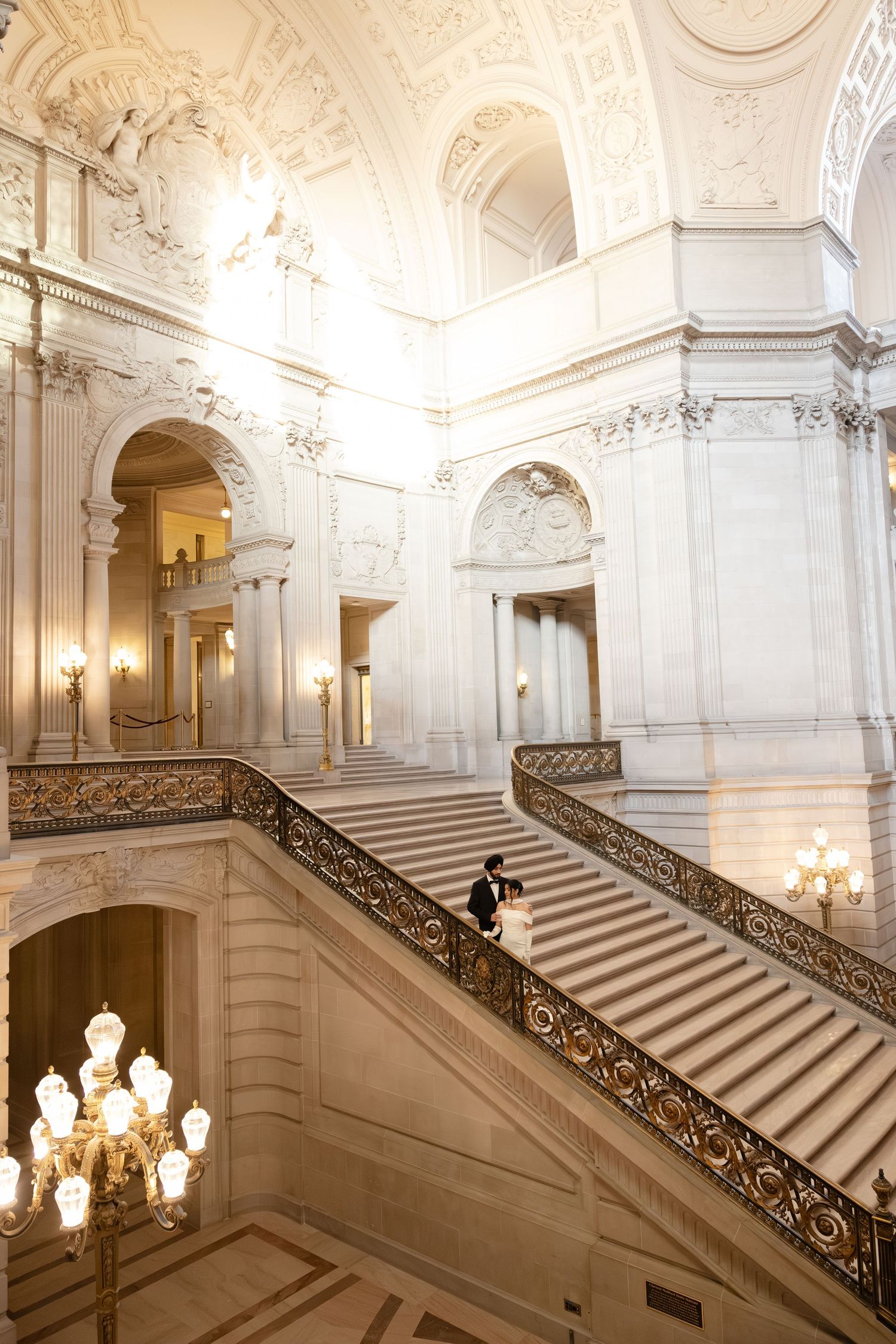Ornate marble staircase with decorative railings and chandeliers inside San Francisco City Hall's grand interior.