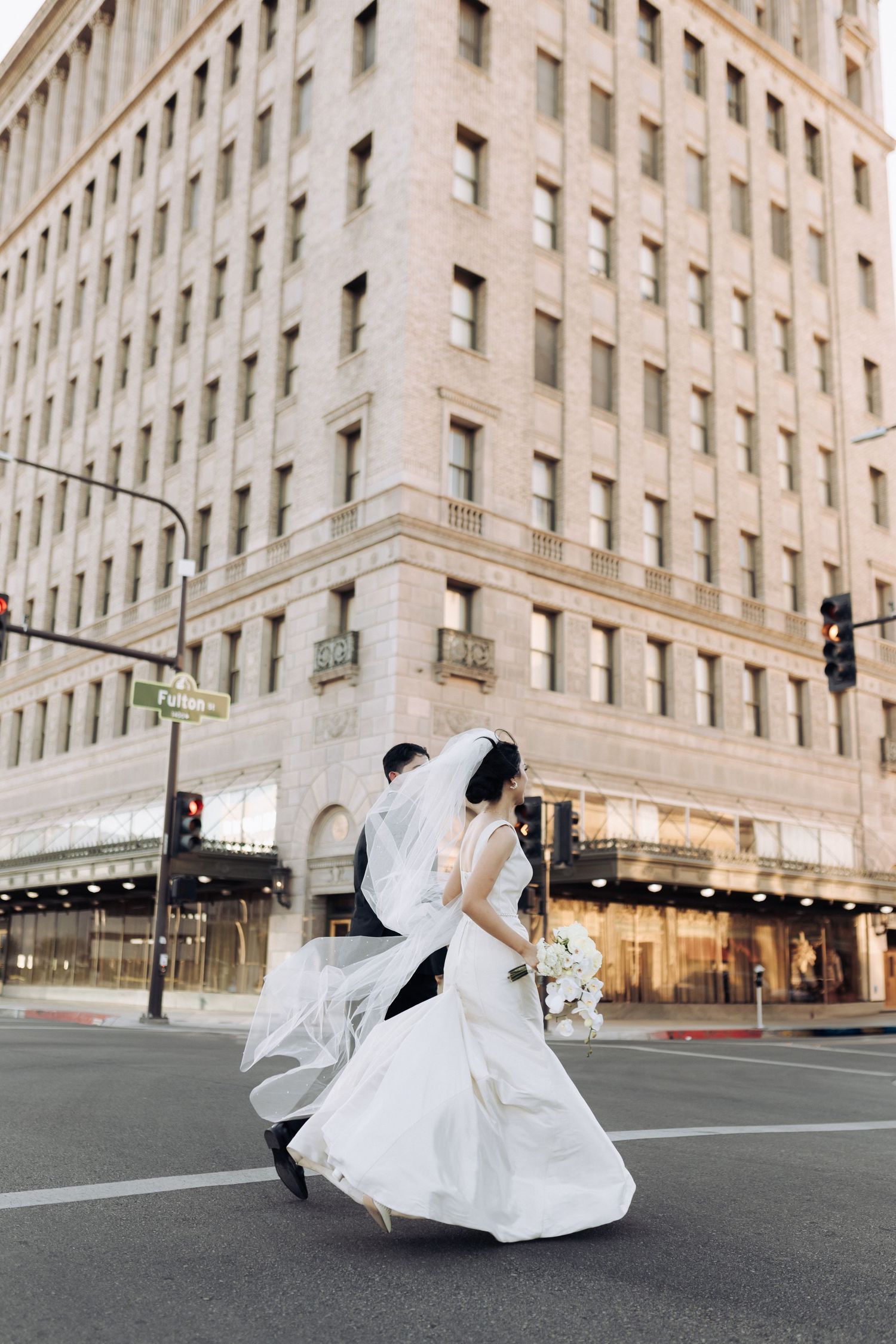 A wedding couple dances and twirls dramatically on an empty city street corner with a historic building backdrop.