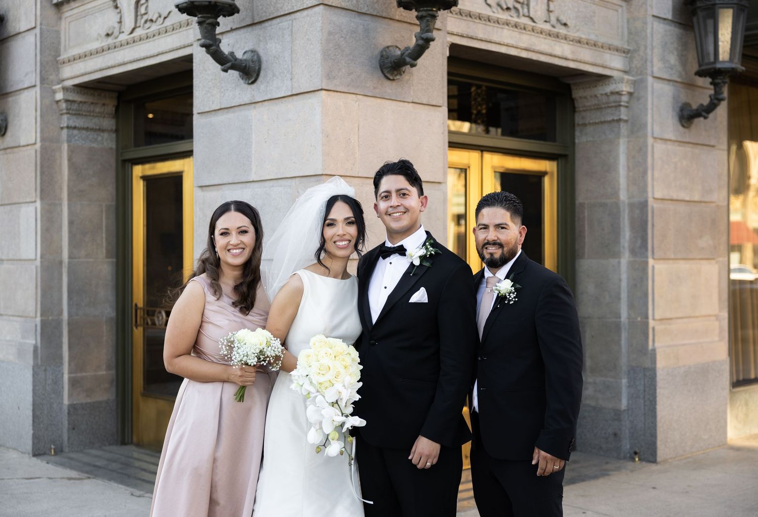 Wedding party members pose outside stone building entrance with ornate lanterns.