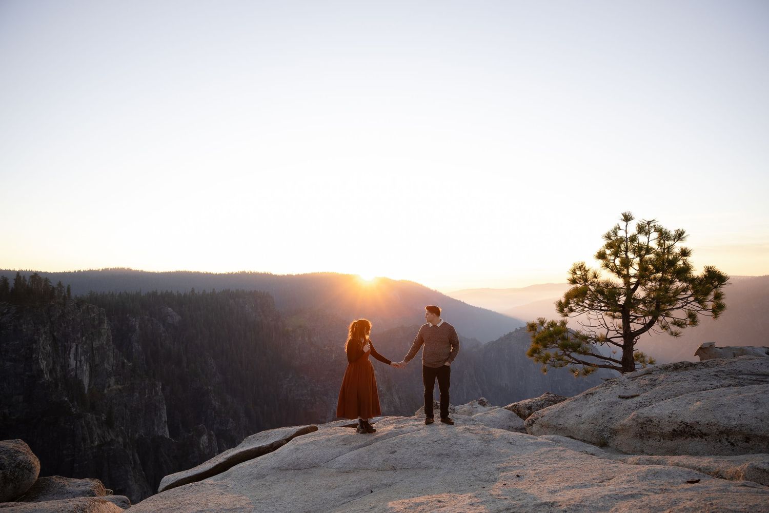 A couple stands on a rocky mountain overlook during sunset in a scenic landscape.