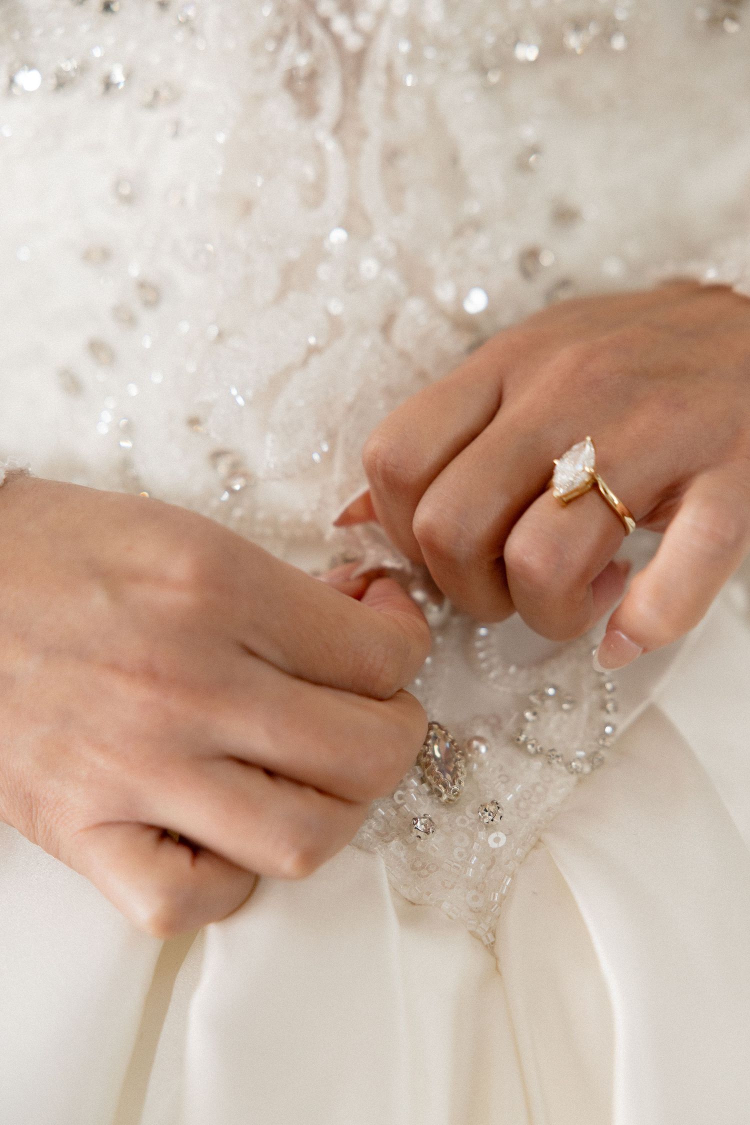 Hands with engagement ring touching ornate beadwork on a white wedding dress.