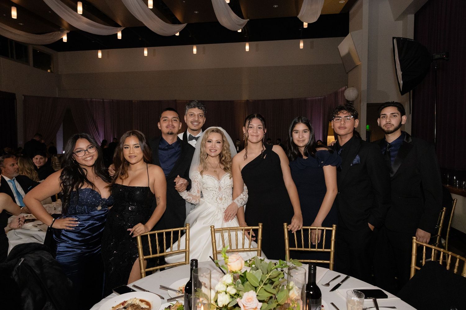 Wedding party gathers around decorated reception table for formal group photo.