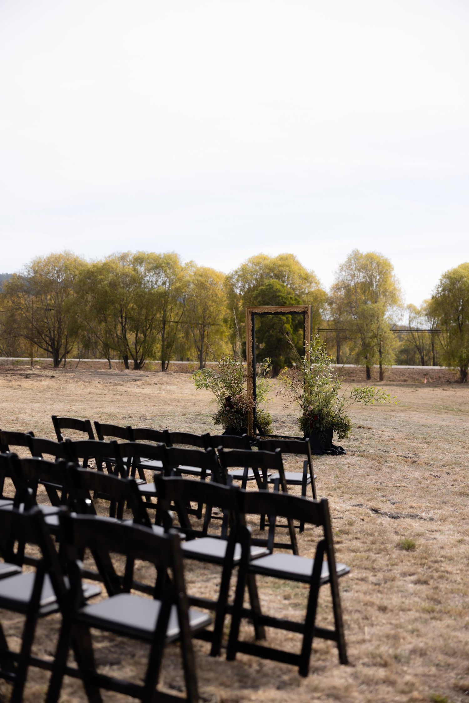 Outdoor wedding ceremony setup with black chairs arranged in rows on dry grass.