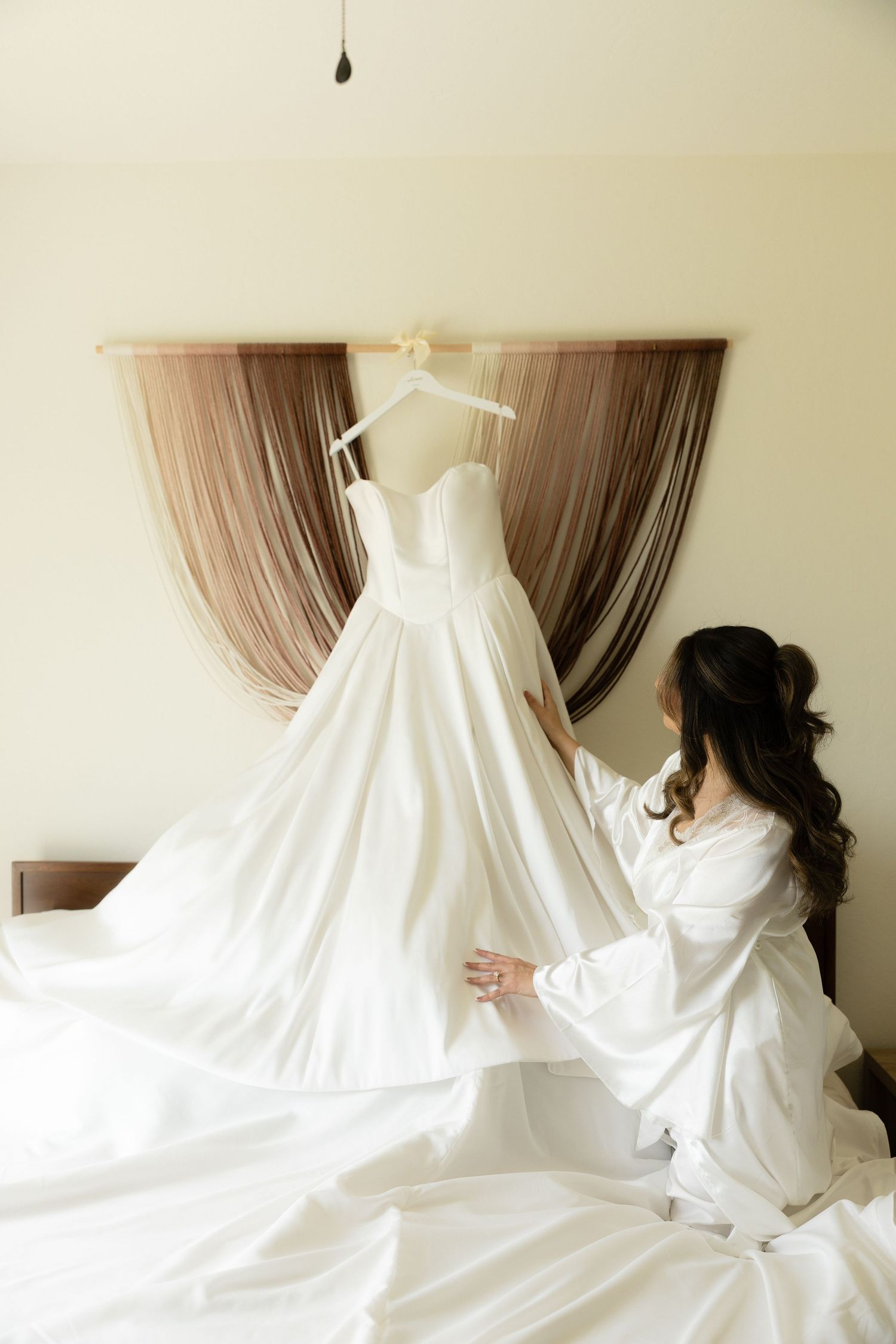 Bride in white robe admiring her strapless ball gown wedding dress hanging above a bed before the ceremony.