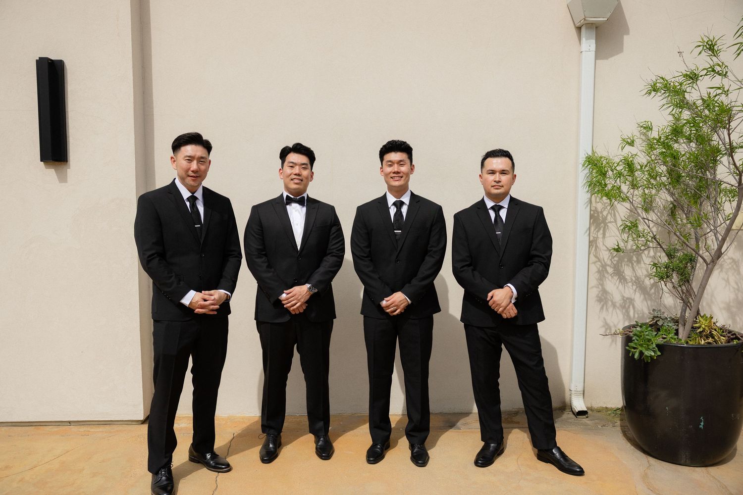 Four groomsmen in matching black suits standing together posing outdoors at a wedding.