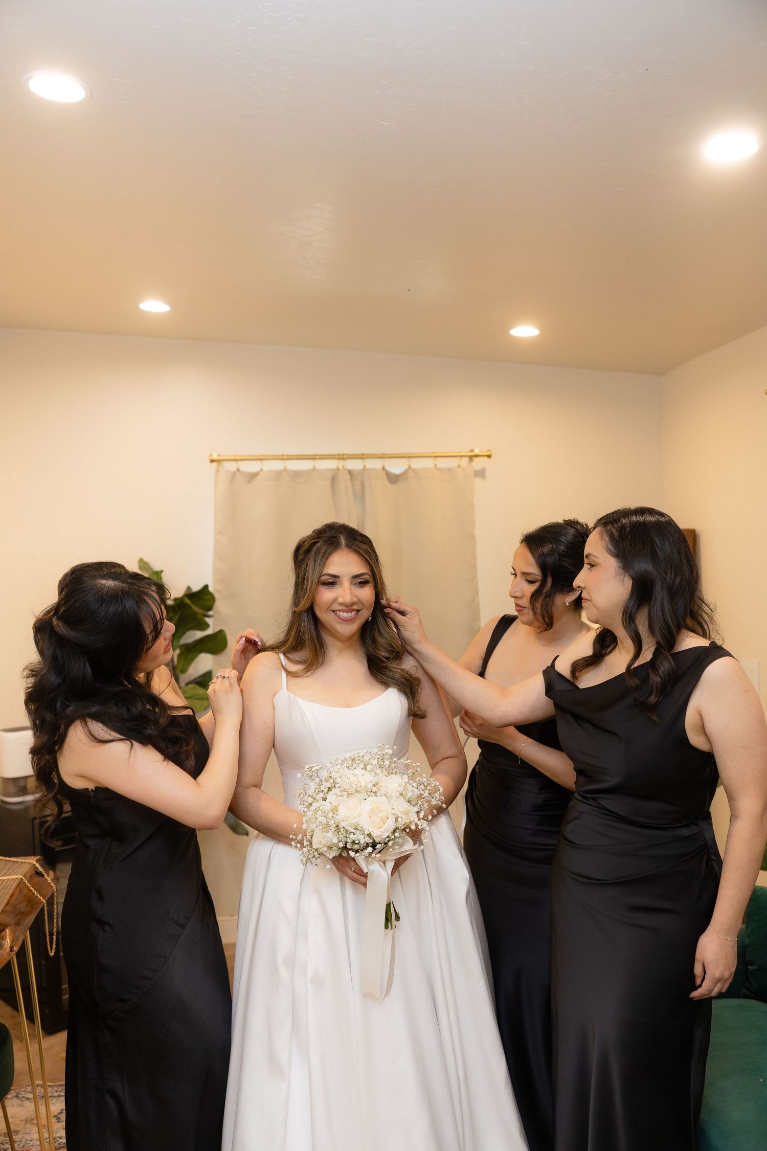 Bride in white gown holds bouquet while bridesmaids in black dresses help her prepare indoors.