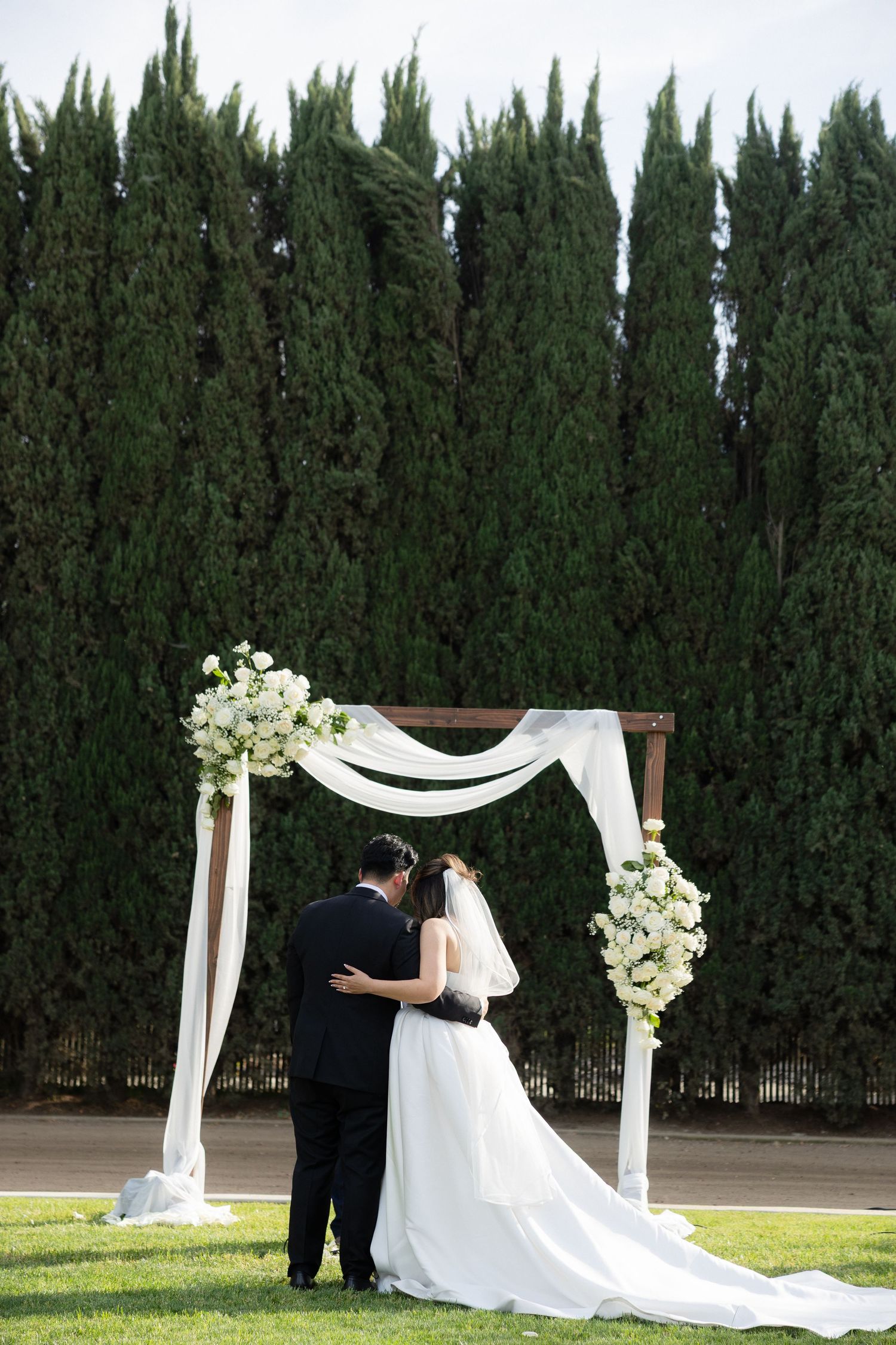 Bride and groom embracing under a white draped floral wedding arch outdoors with tall cypress trees backdrop.