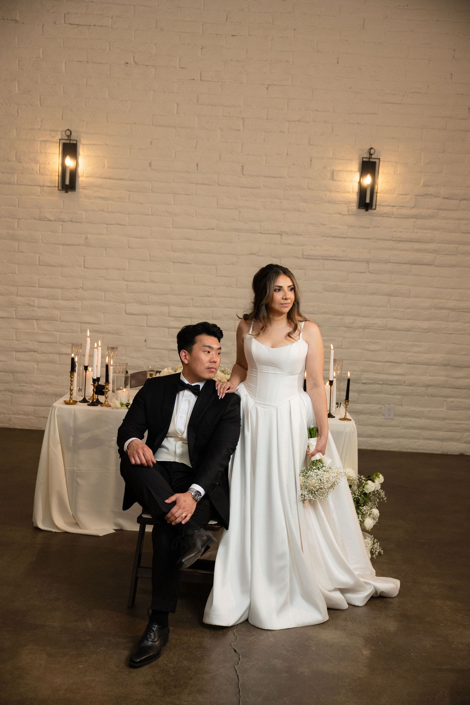 Wedding couple poses before sweetheart table with candles, groom seated in black tux, bride standing in white gown.