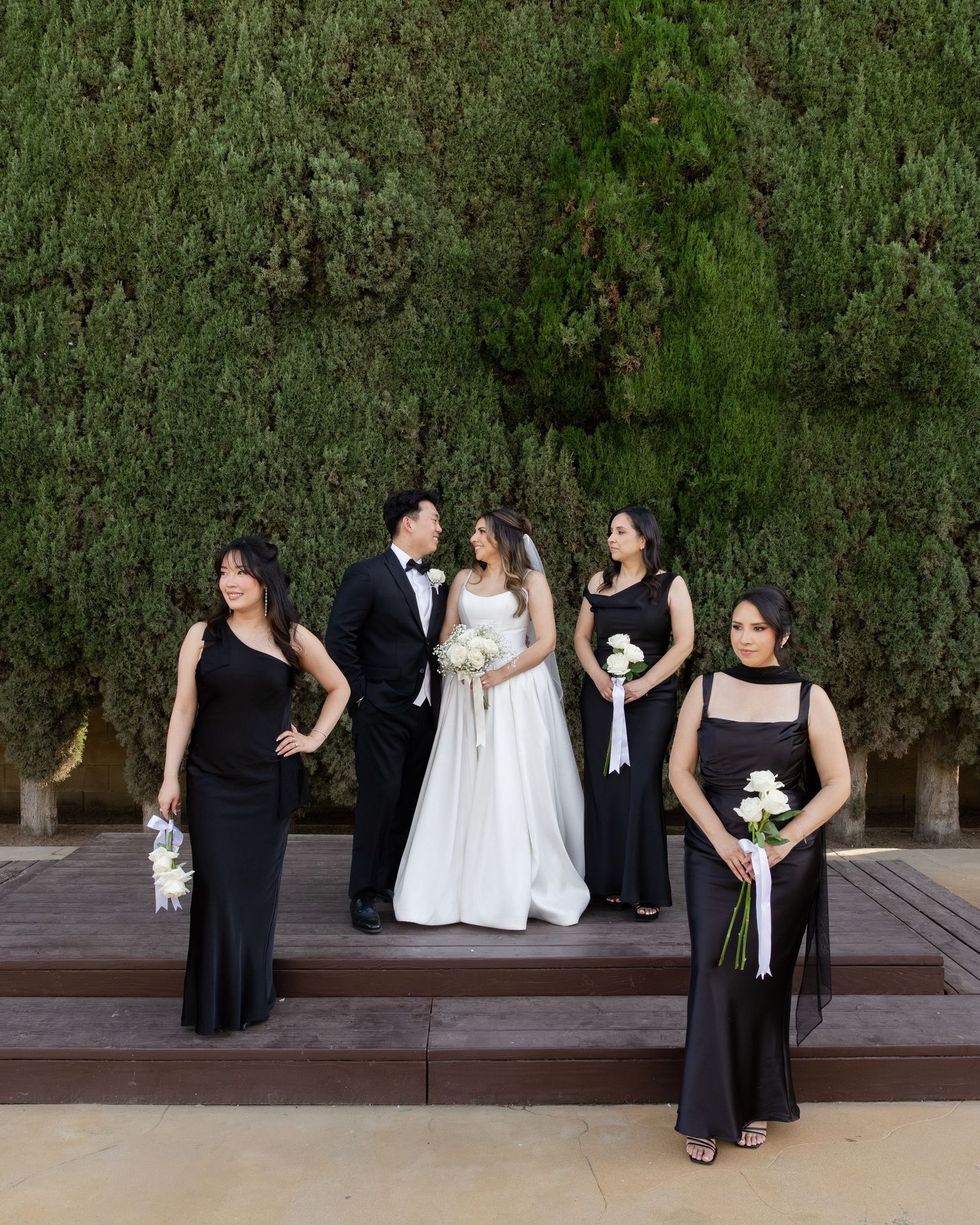 Bride and groom share a romantic moment with bridesmaids in black gowns holding white bouquets outdoors.