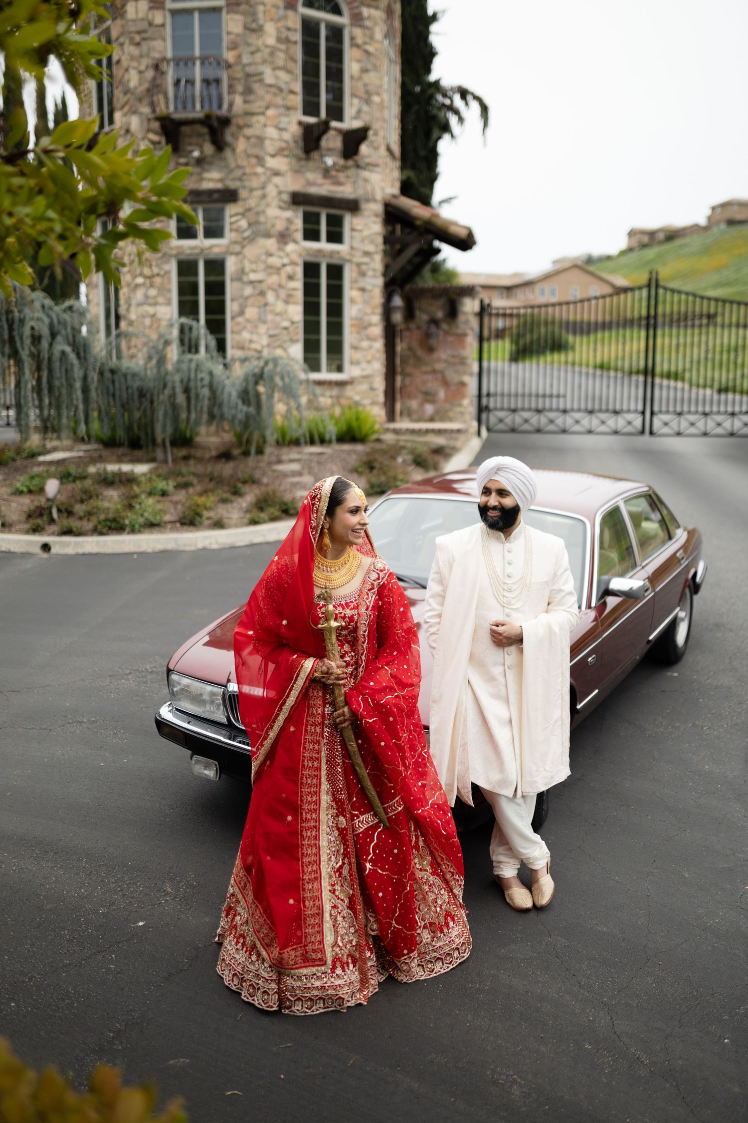 A couple in traditional South Asian wedding attire pose together near a vintage car outside a stone building.