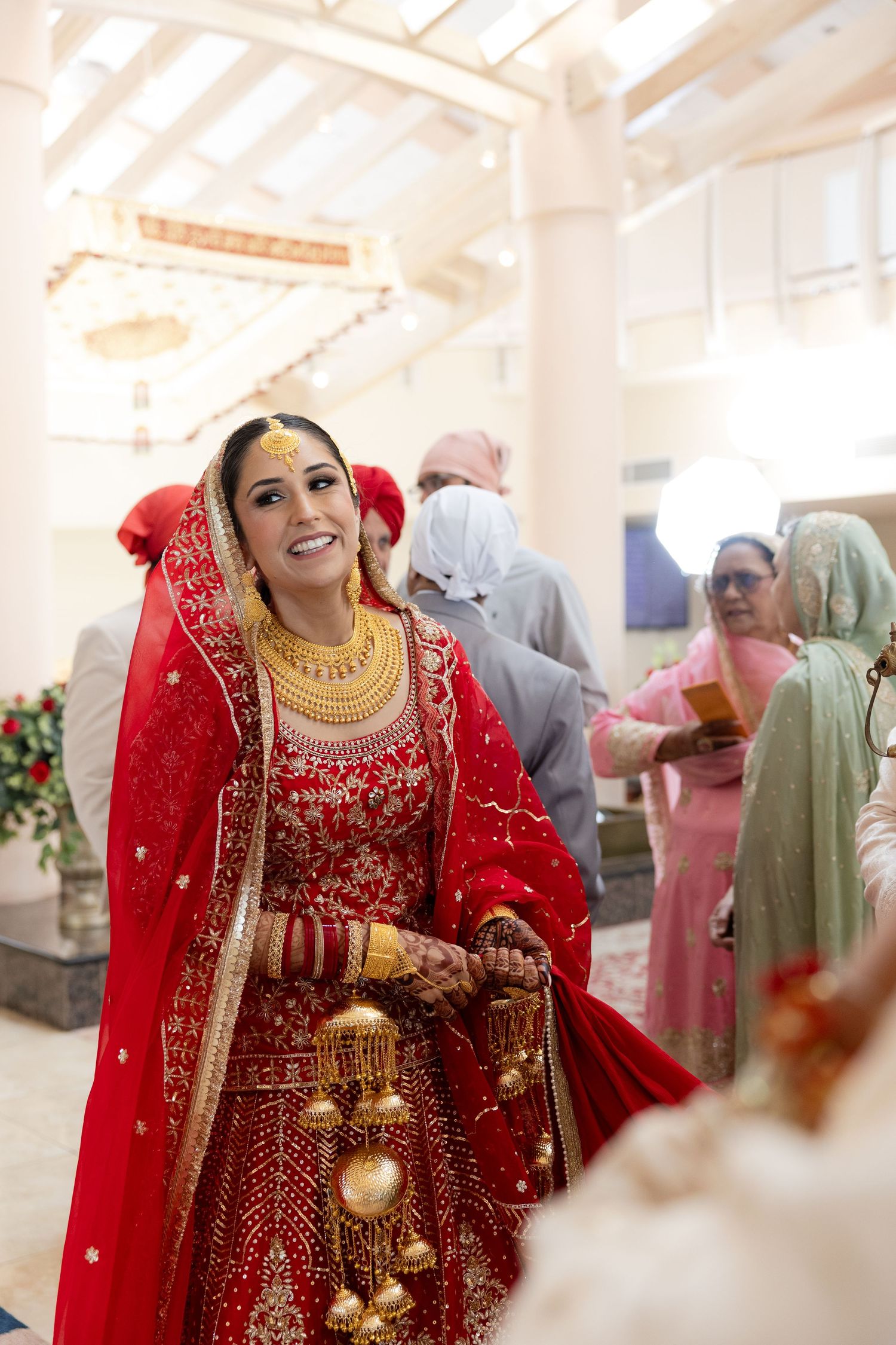 A bride in a traditional red Indian wedding lehenga with gold jewelry smiles during her celebration.