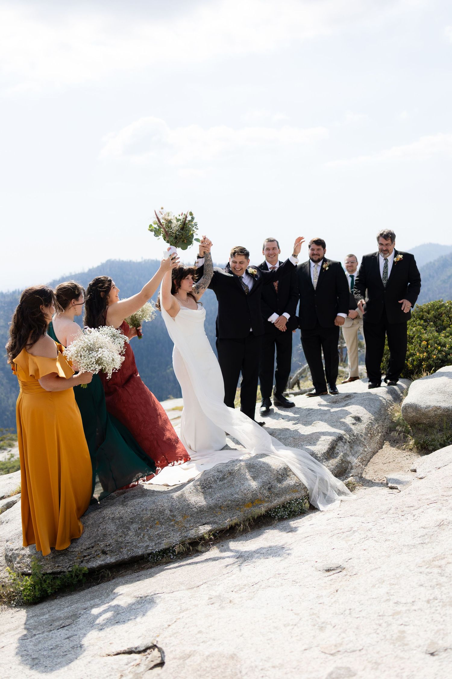 A joyful wedding party celebrates on a rocky mountain summit, with the bride and groom raising their arms in triumph.
