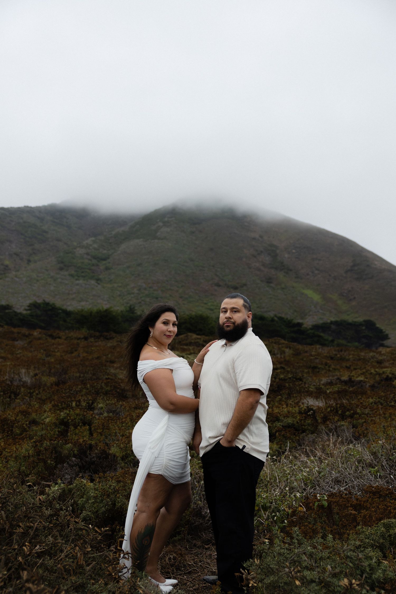 Couple posing together in white outfits on a misty hillside with green shrubs and foggy mountain backdrop.