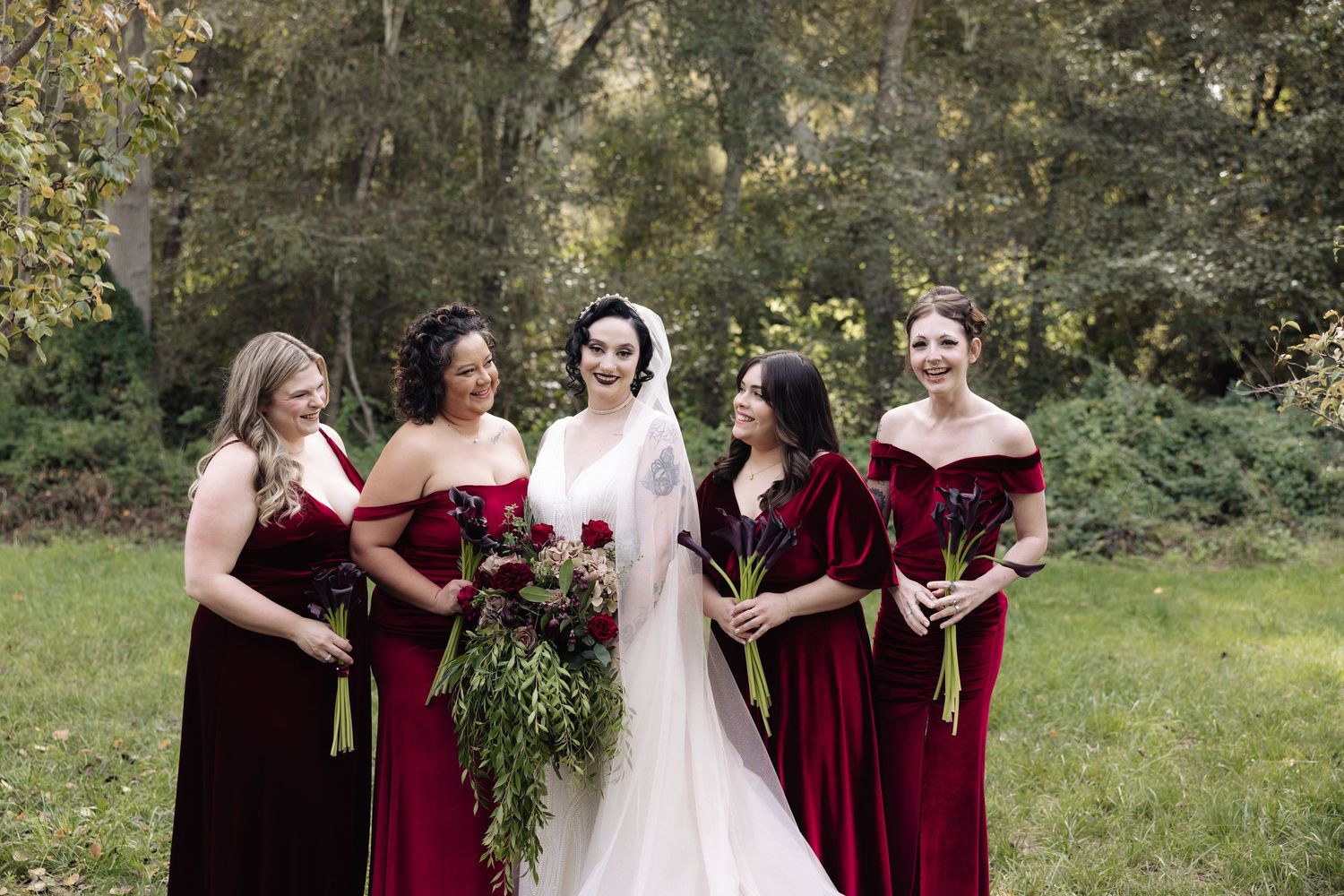 Wedding party in burgundy dresses and black suits poses playfully in an outdoor garden setting.