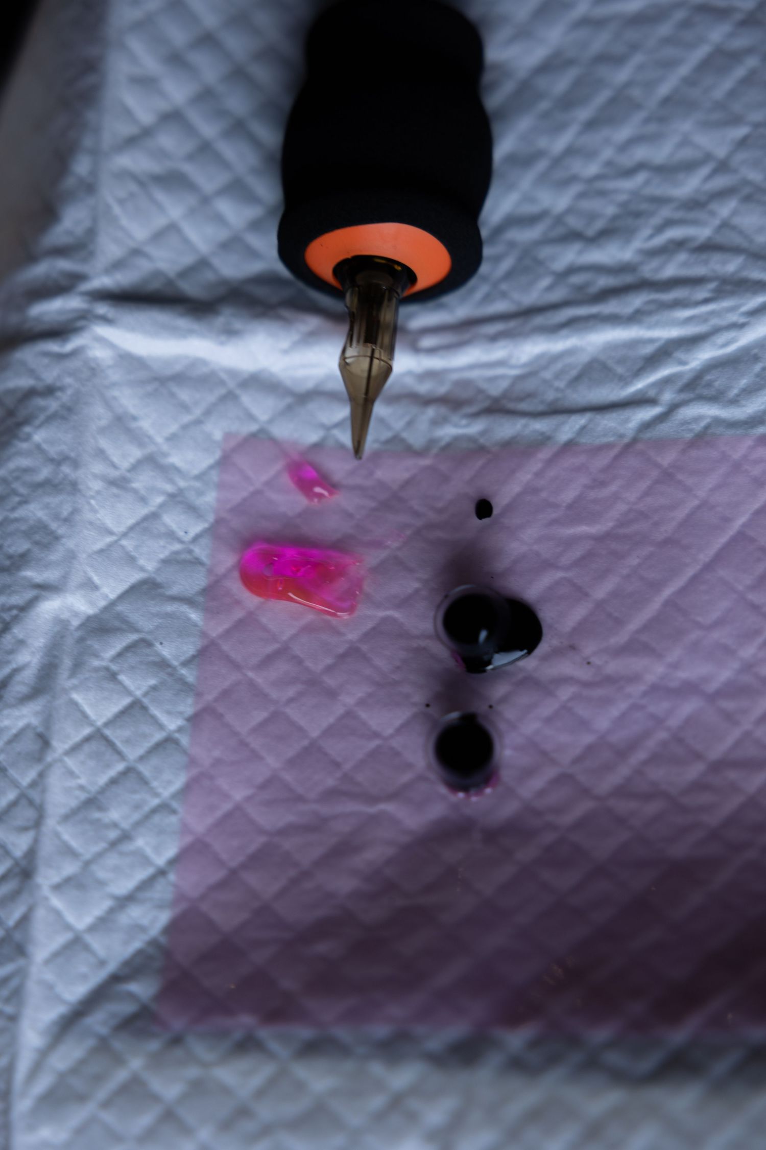 Close-up of calligraphy pen with pink and purple ink drops on white quilted surface.