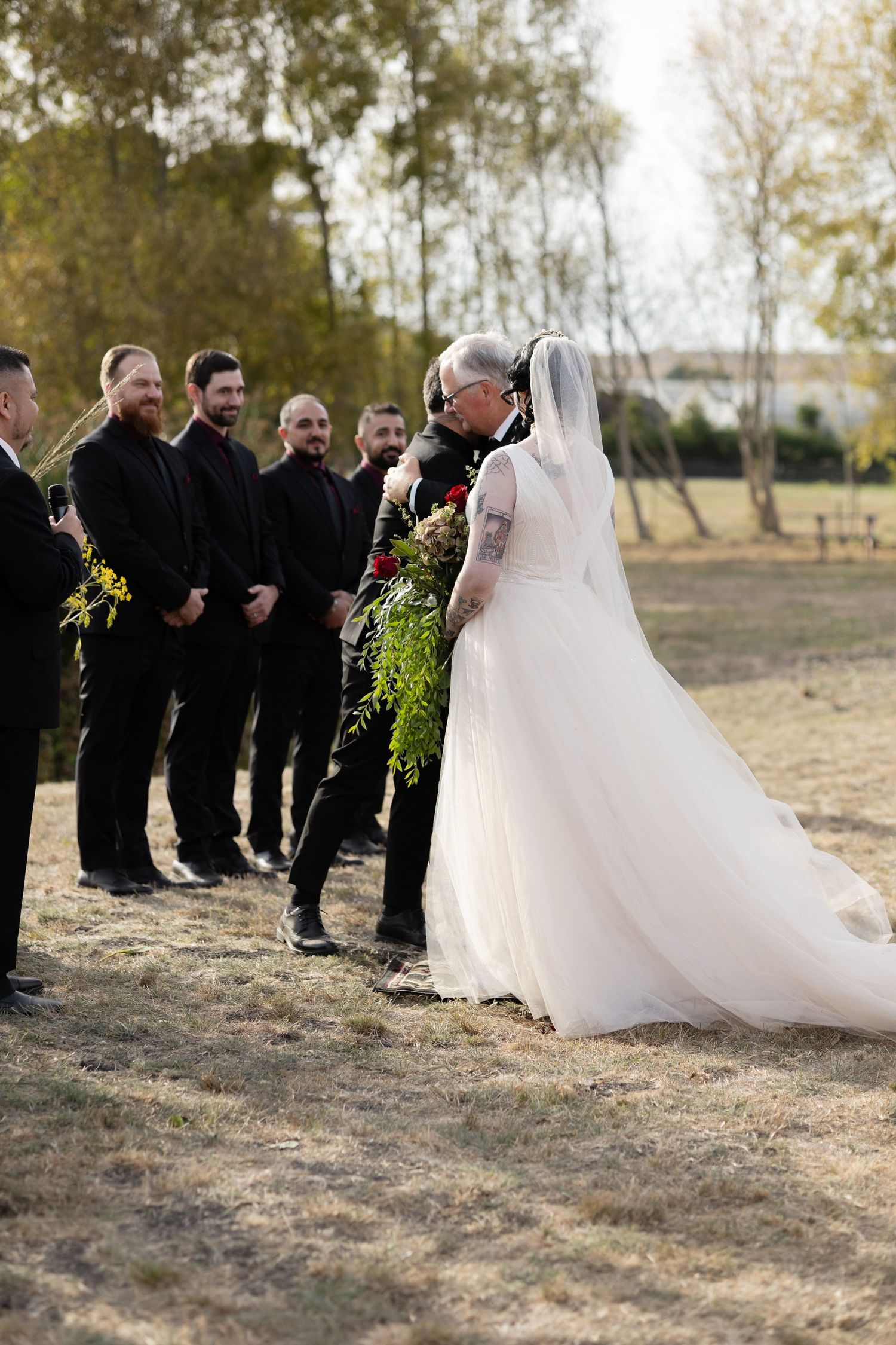 Wedding party standing in formation on dry grass with trees in background.