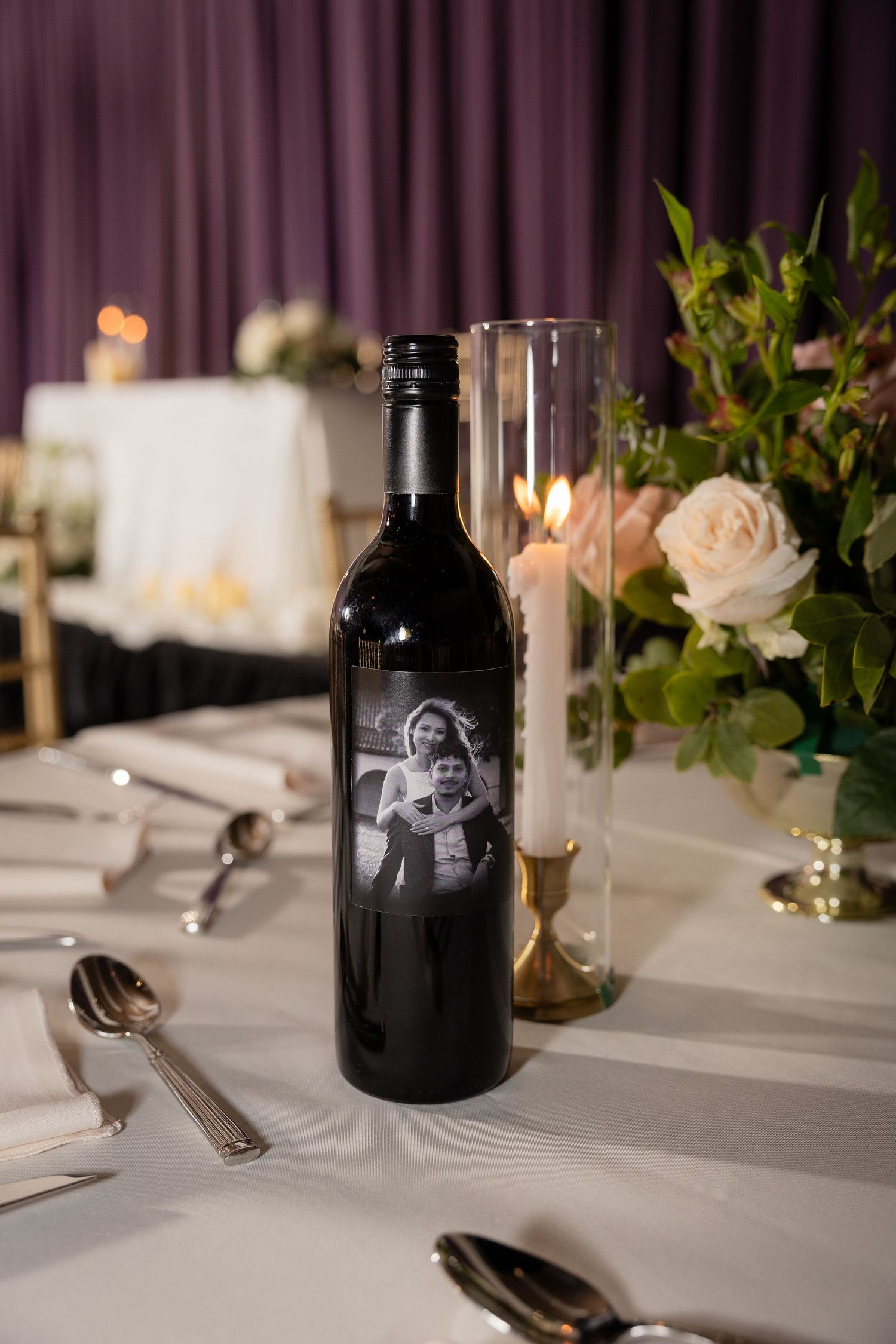 A custom wine bottle with a black and white photo sits on an elegant wedding reception table with roses and candles.