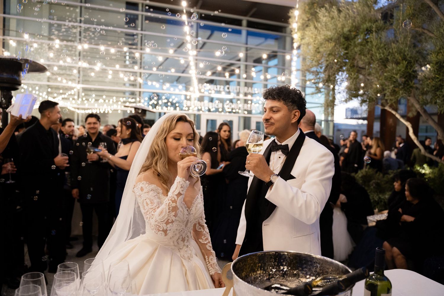 A bride and groom celebrate with guests during their wedding reception under twinkling lights.