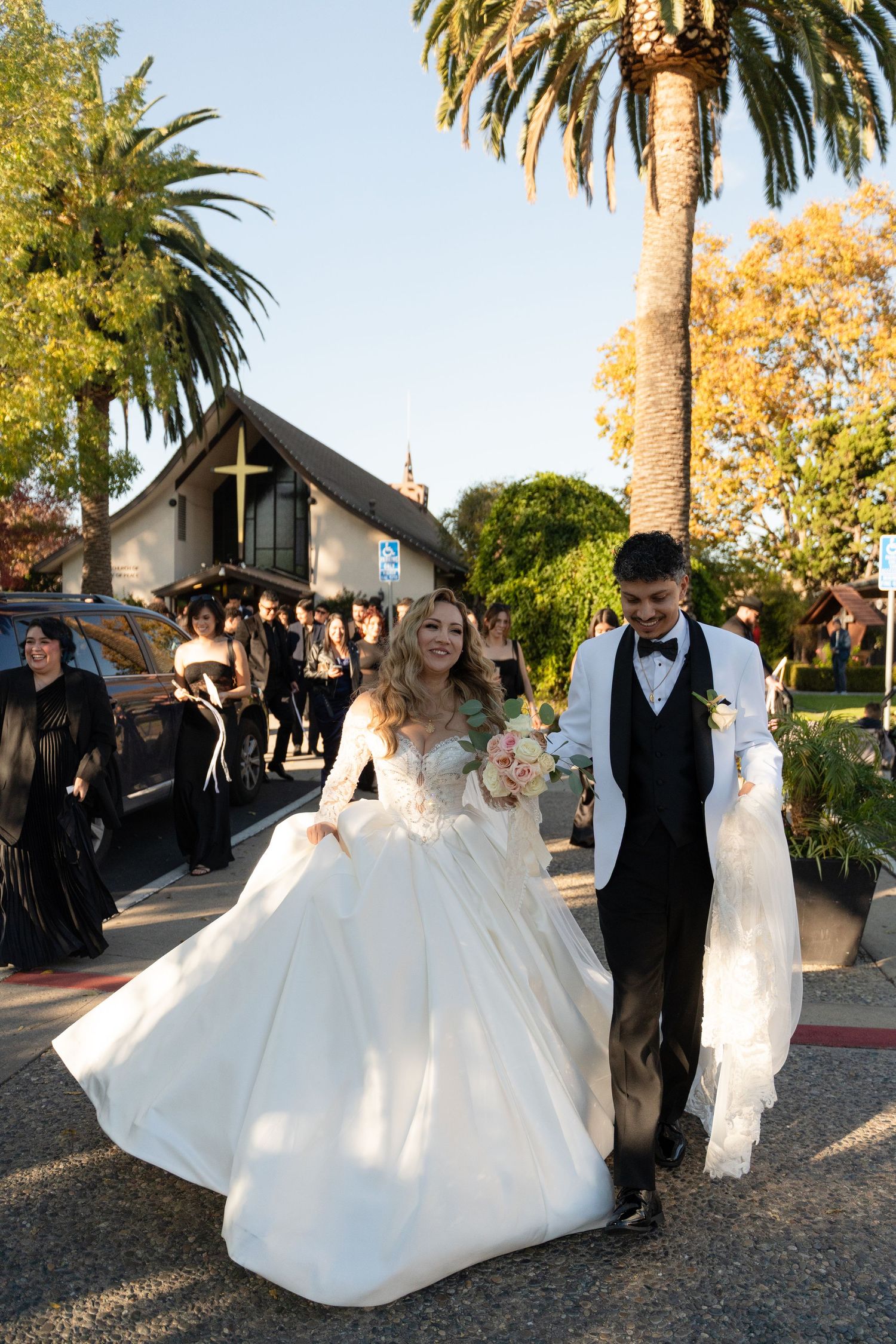Wedding couple walks outside a church with palm trees and guests in black attire.