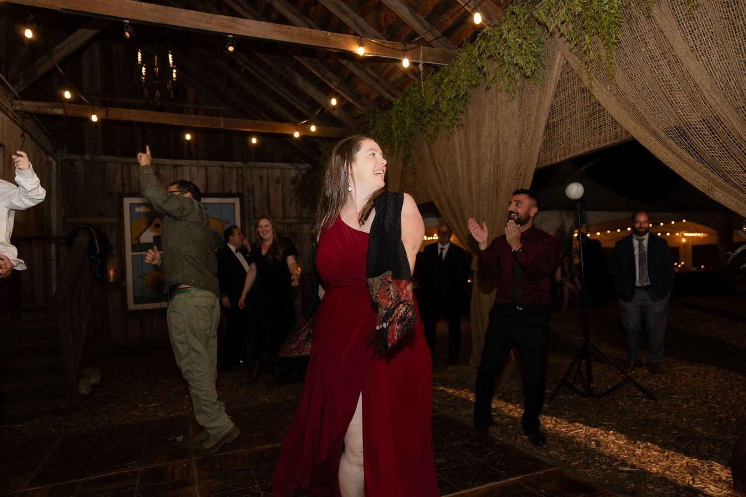 People dancing and celebrating at a rustic barn wedding reception with string lights and wooden beams overhead.