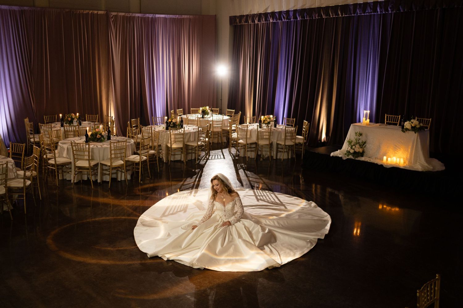 A dramatic wedding dress train flows across a dark ballroom floor with elegant lighting and table settings.