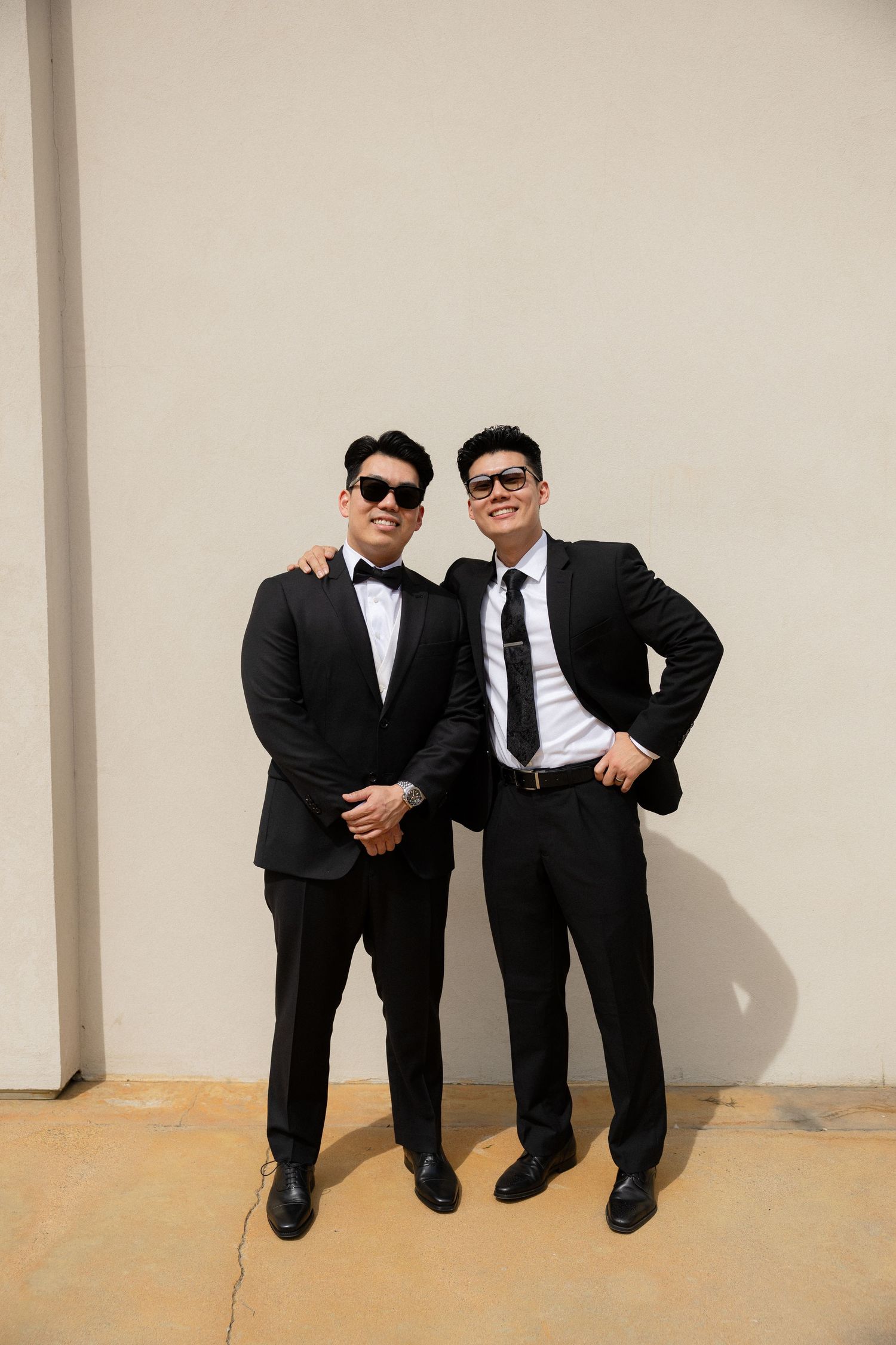 Two groomsmen wearing black suits and sunglasses posing confidently against a white wall at a wedding.