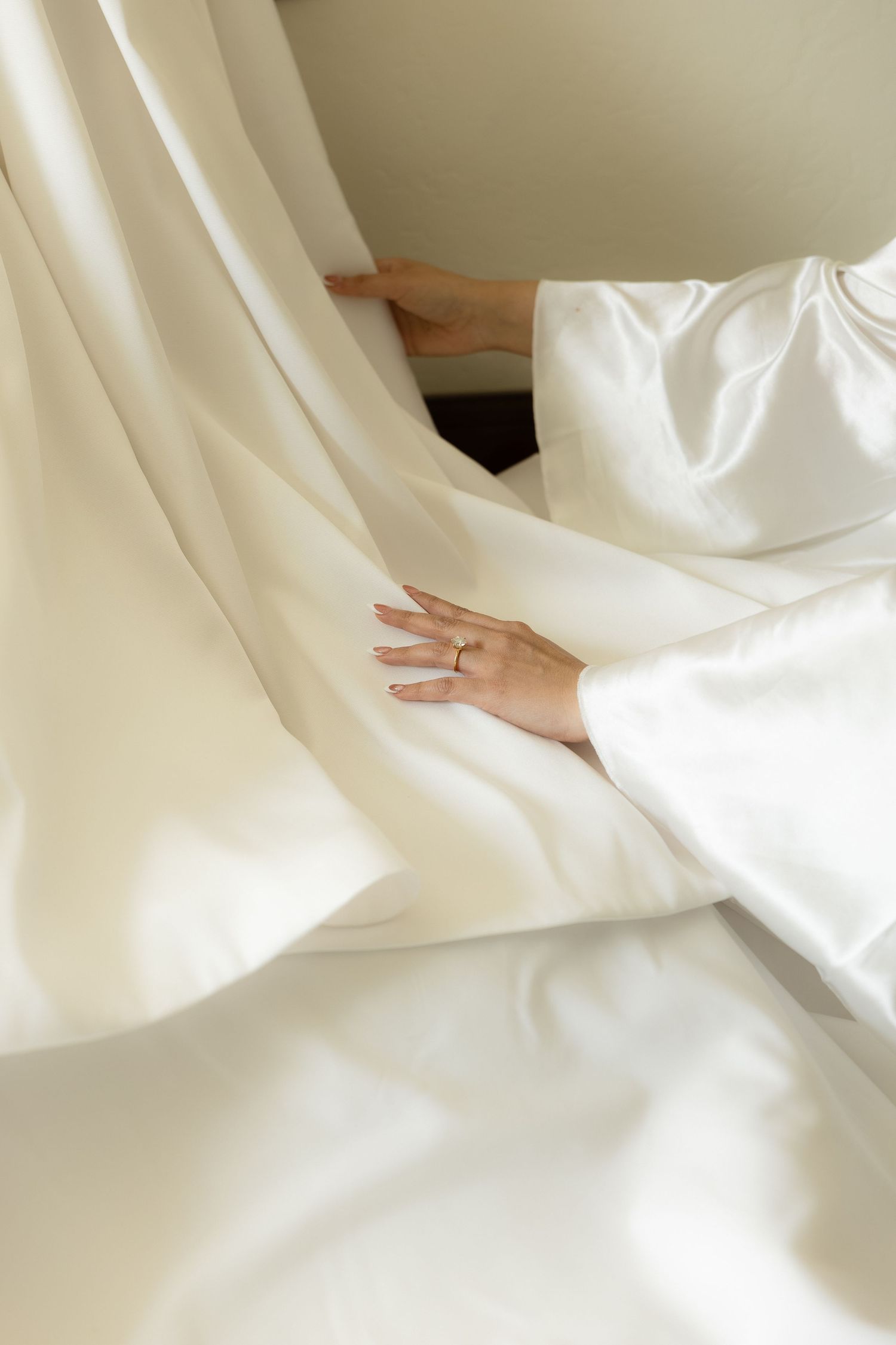 Close-up of a bride's hand with engagement ring resting on white satin wedding dress fabric.