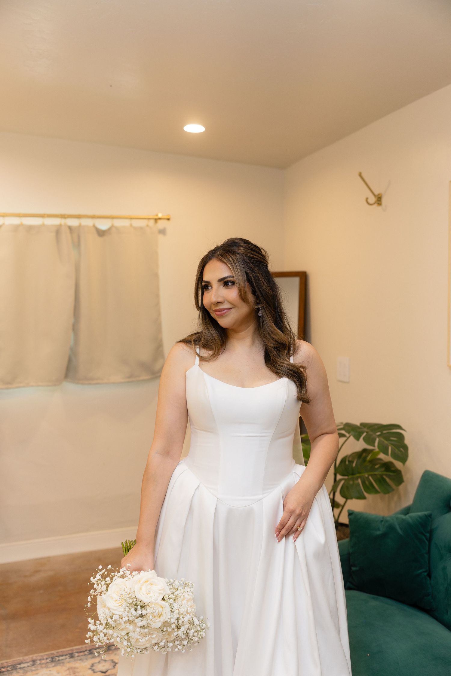 Bride in elegant white satin wedding gown holding white floral bouquet standing in bridal suite.
