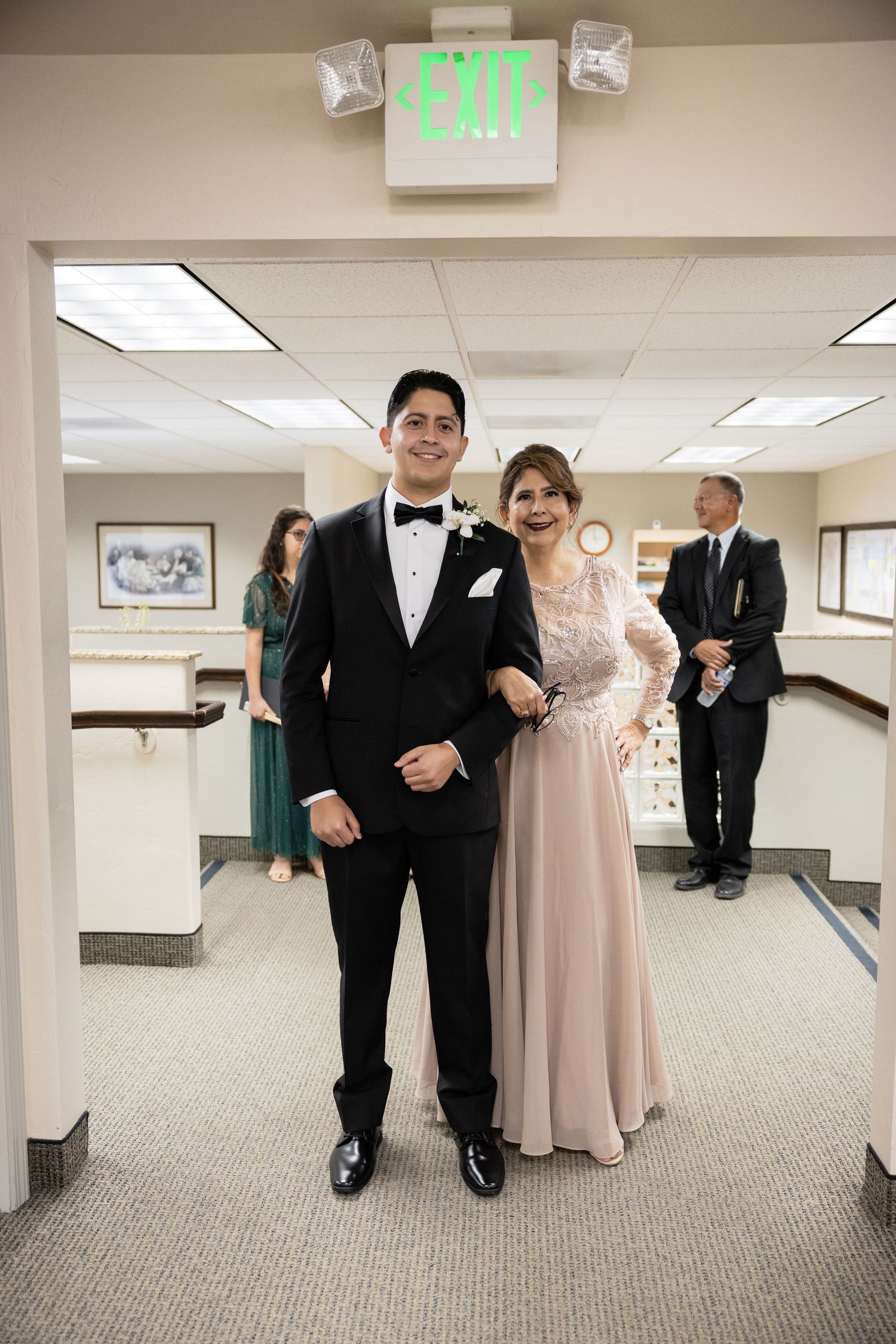Wedding party members walking through a hallway with fluorescent lighting and exit signs.