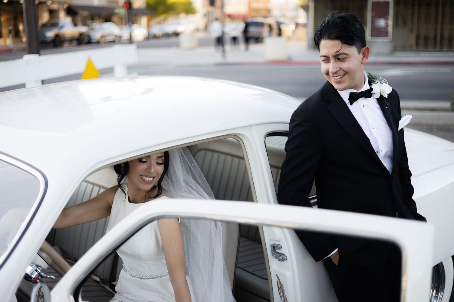 Elegant wedding couple posing with a pristine white vintage car against an urban backdrop.