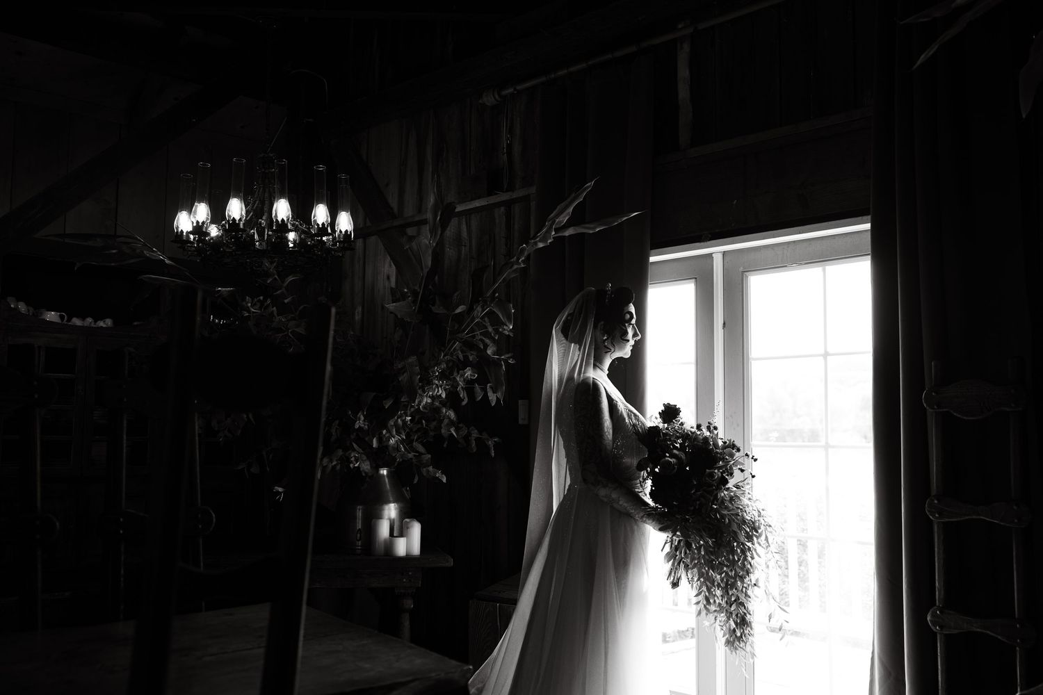 Black and white photo of a bride in silhouette holding flowers by a window with chandelier.