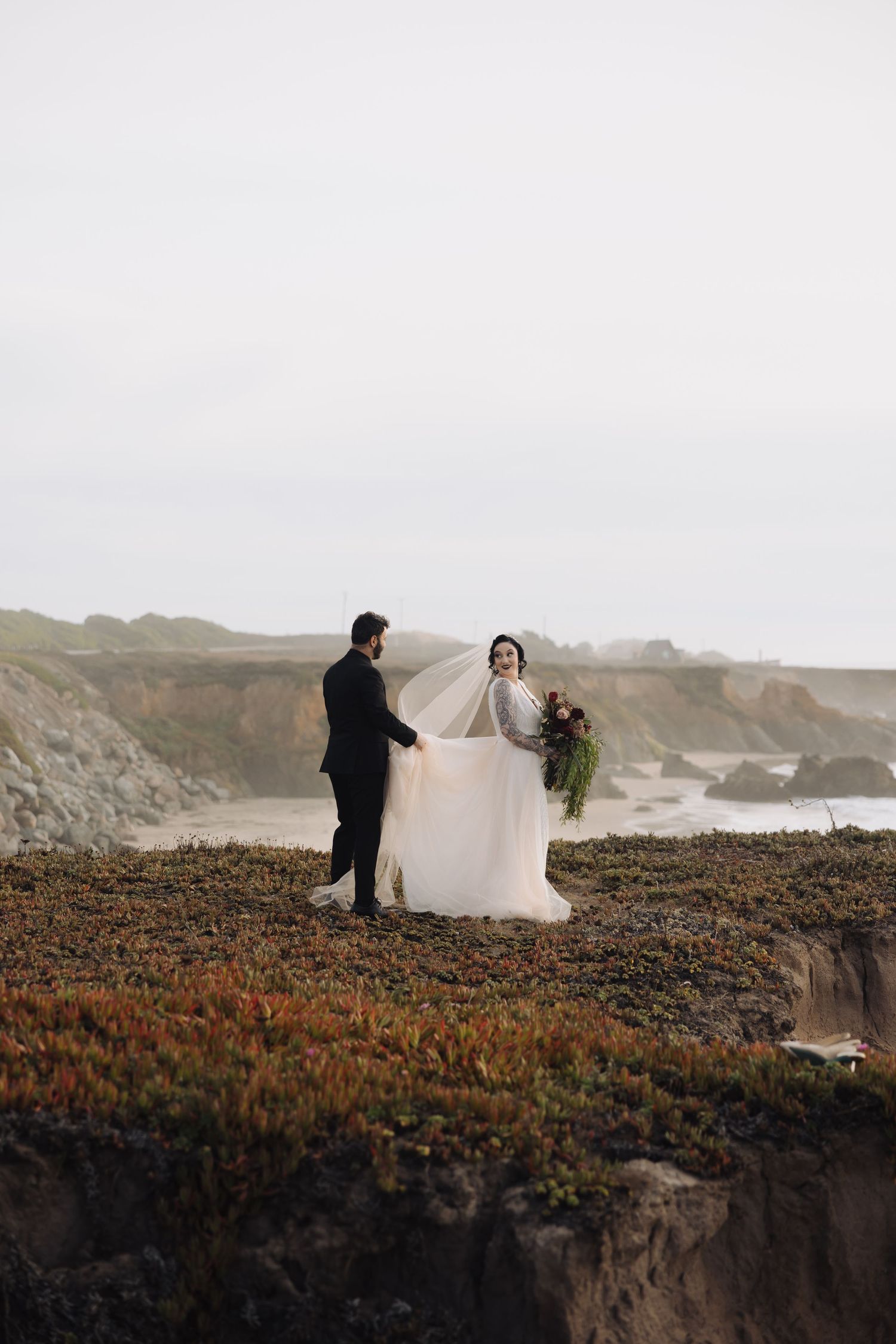 A wedding couple poses on windswept coastal cliffs with dramatic ocean views.