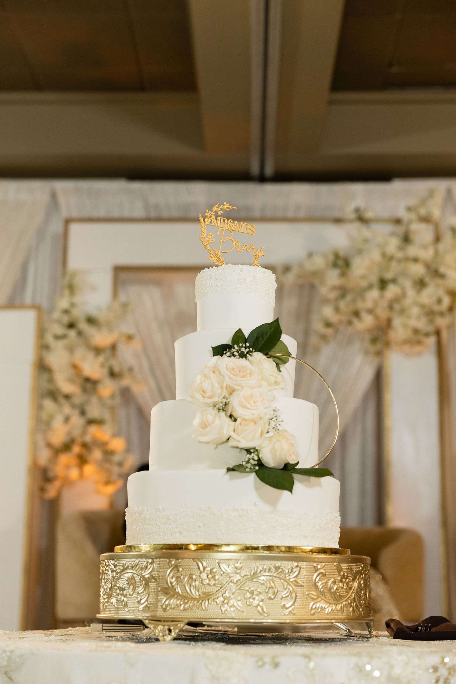 Three-tiered white wedding cake decorated with fresh flowers sits atop an ornate gold cake stand.