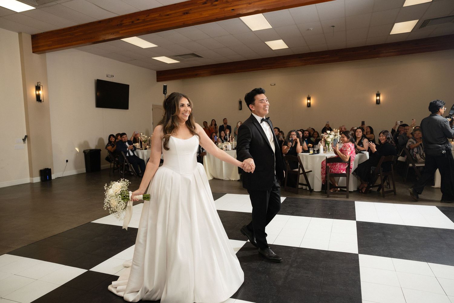 Bride in strapless white ball gown and groom in black tuxedo hold hands entering wedding reception on checkered dance floor.