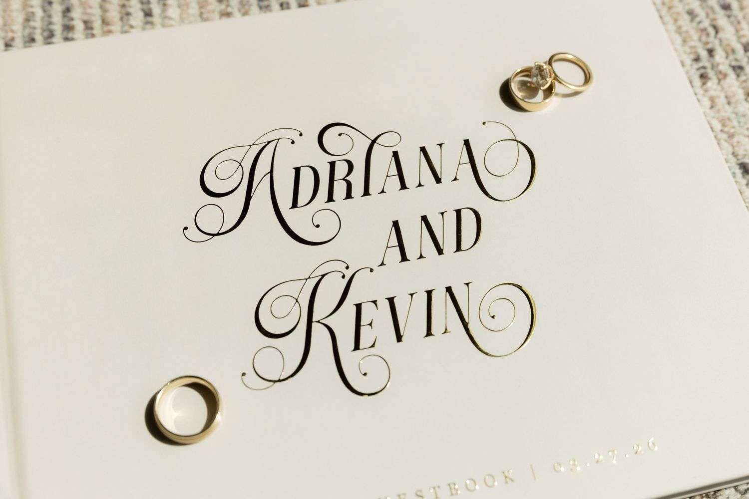 Elegant wedding stationery with ornate calligraphy reading Adriana and Kevin, adorned with gold wedding rings on white background.
