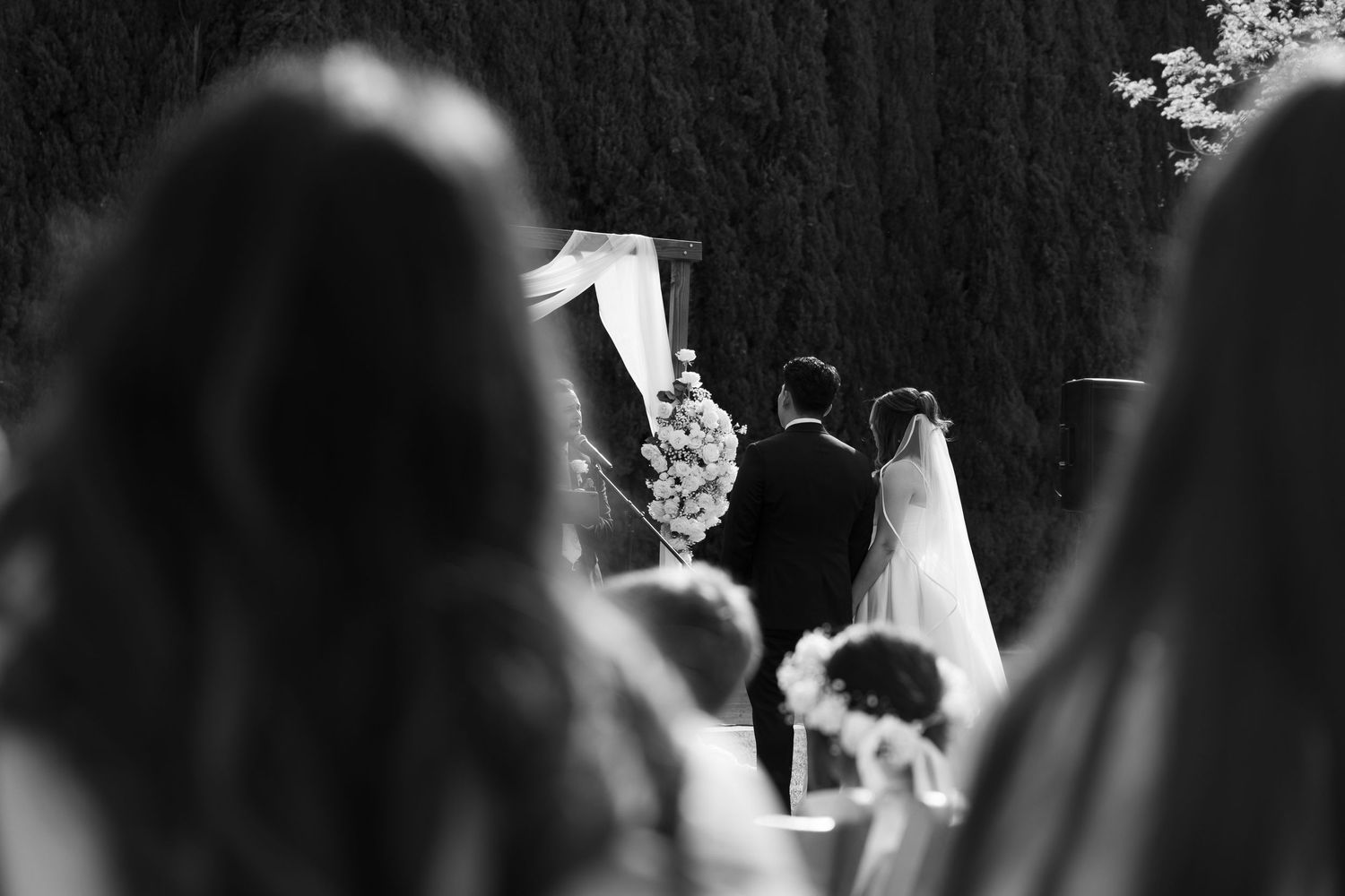 Black and white photo of wedding guests watching couple exchange vows at floral altar.