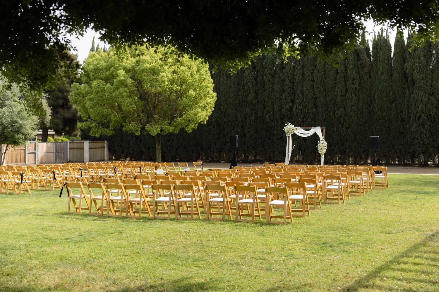 Outdoor wedding ceremony setup with rows of wooden folding chairs on green lawn, floral arch, and tall cypress trees backdrop.
