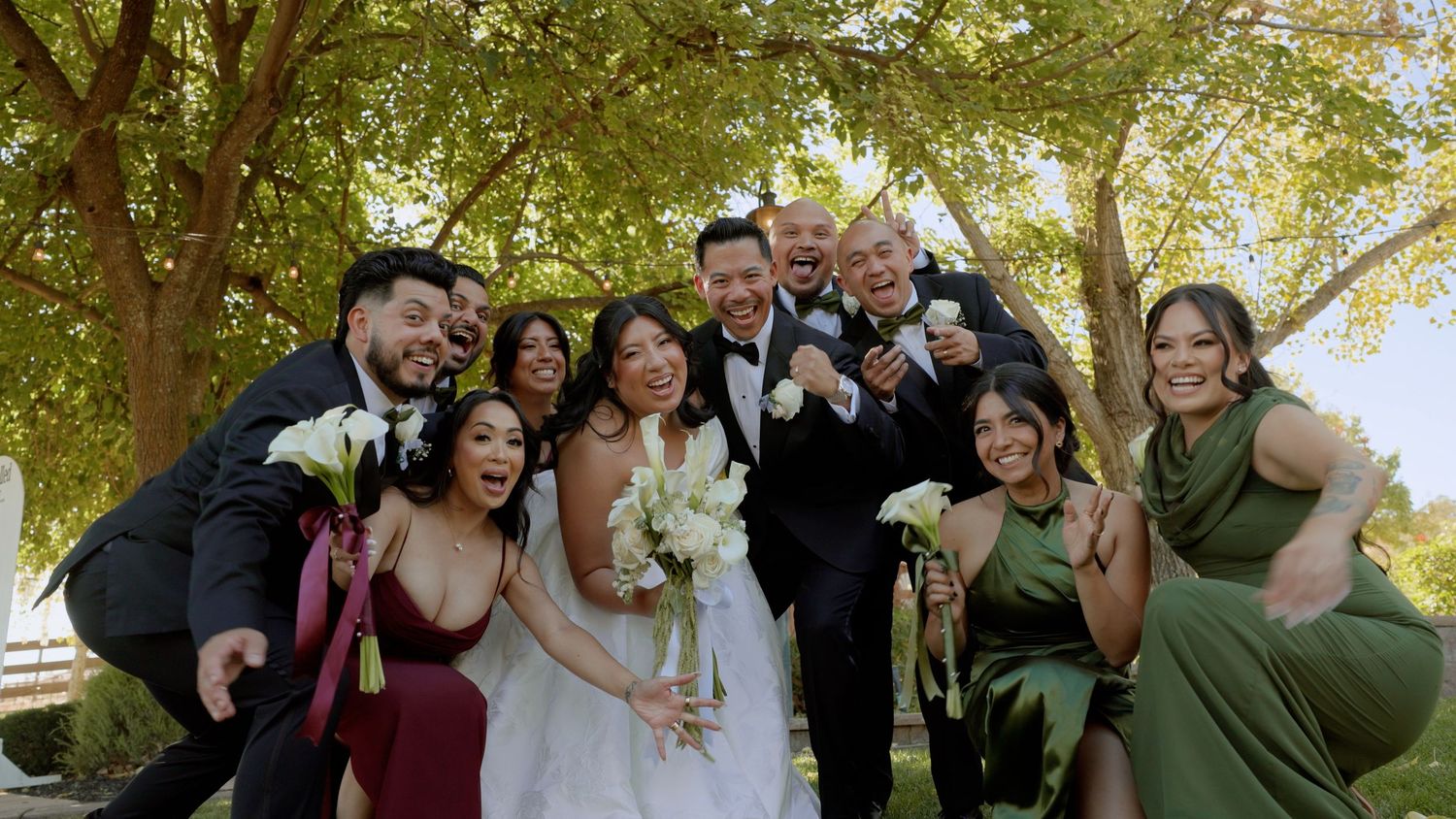 Wedding party poses joyfully outdoors under trees, bride in white gown with bouquet surrounded by groomsmen and bridesmaids in green.